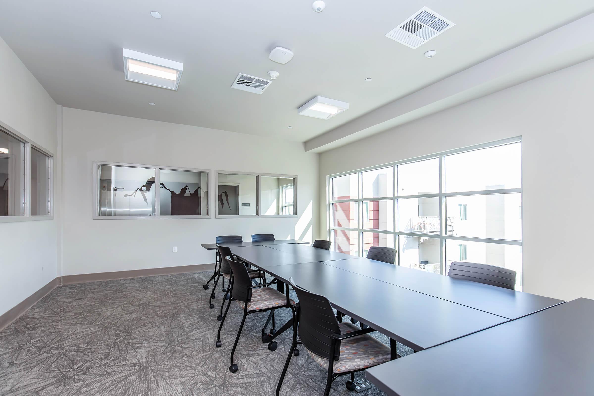 A modern conference room featuring a long rectangular table surrounded by several black chairs. Large windows allow natural light to fill the space, and there are lights on the ceiling. A glass partition on one wall showcases a separate area. The flooring has a textured pattern, contributing to the contemporary design.