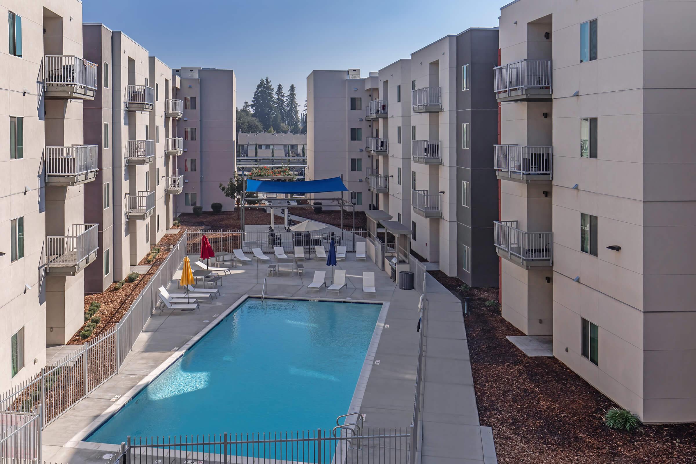 A view of a residential complex featuring a central swimming pool surrounded by lounge chairs, umbrellas, and landscaped areas. The surrounding buildings are modern with balconies and large windows, situated in a well-lit, sunny environment.