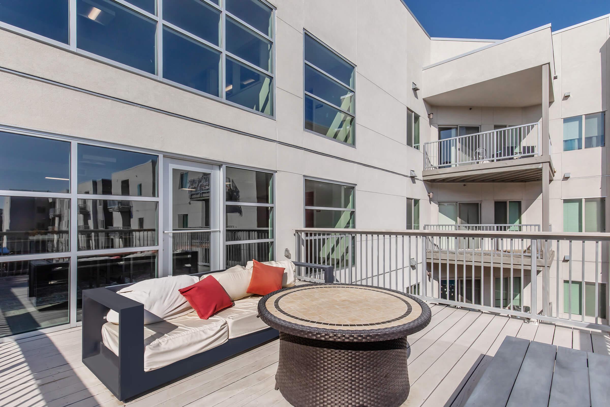 A modern outdoor balcony with a comfortable sofa featuring red accent pillows, a round table, and large windows of an apartment building in the background. The space is bright and airy, with clear blue skies above and a wooden deck.