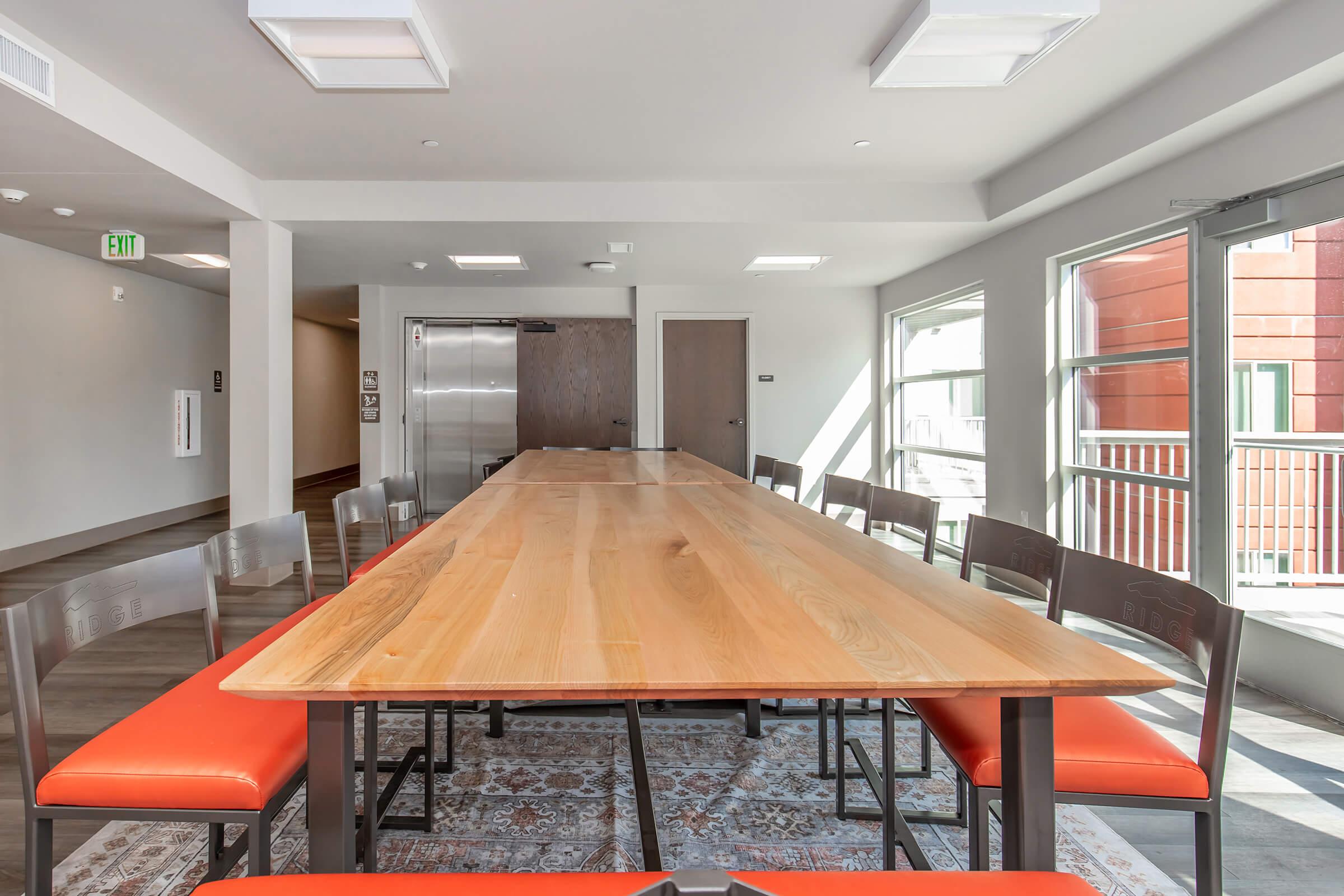 A spacious, modern meeting room featuring a long wooden table surrounded by chairs with bright orange cushions. Large windows allow natural light, and the floor has a patterned rug. An elevator is visible in the background, contributing to the contemporary atmosphere of the space.