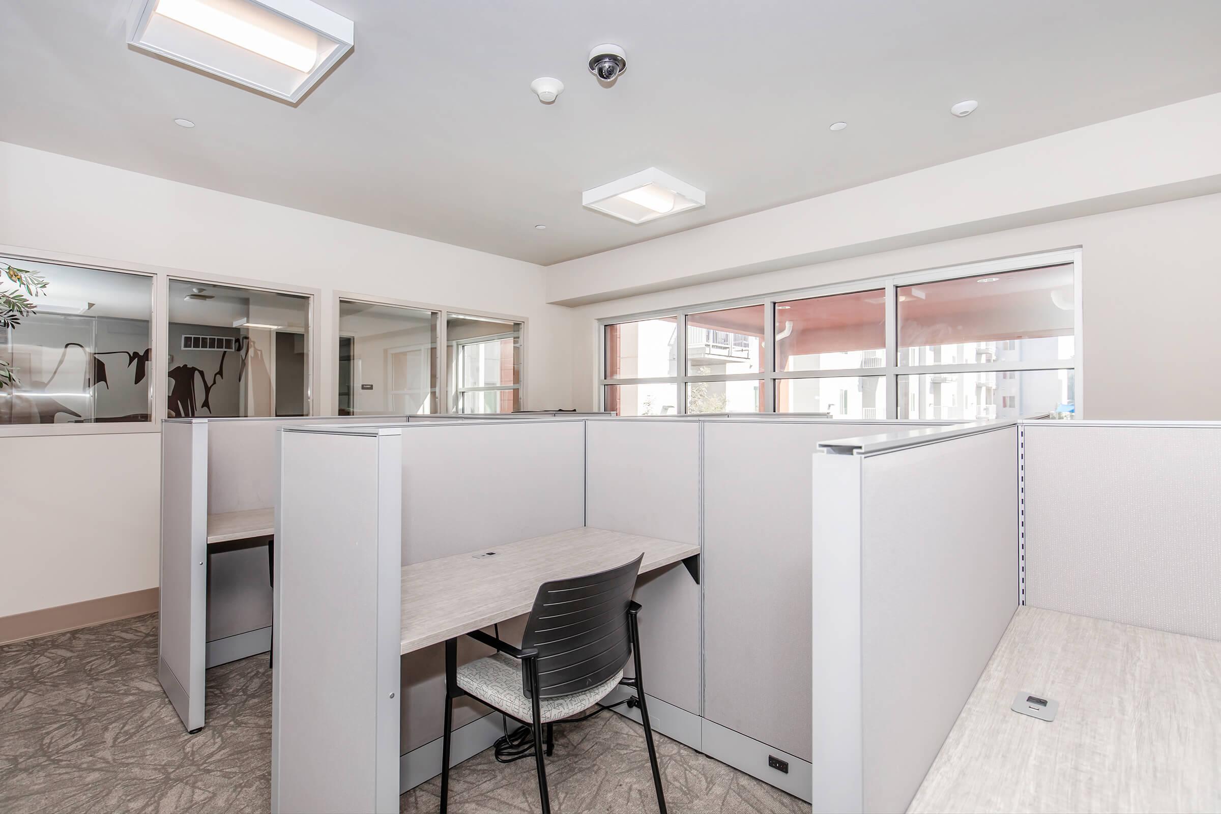 A modern office space featuring several cubicles with light-colored partitions. Each cubicle has a simple desk and a chair, with natural light coming through large windows. The walls are painted in a soft neutral color, creating a calm work environment. A ceiling-mounted camera is visible, indicating security measures.