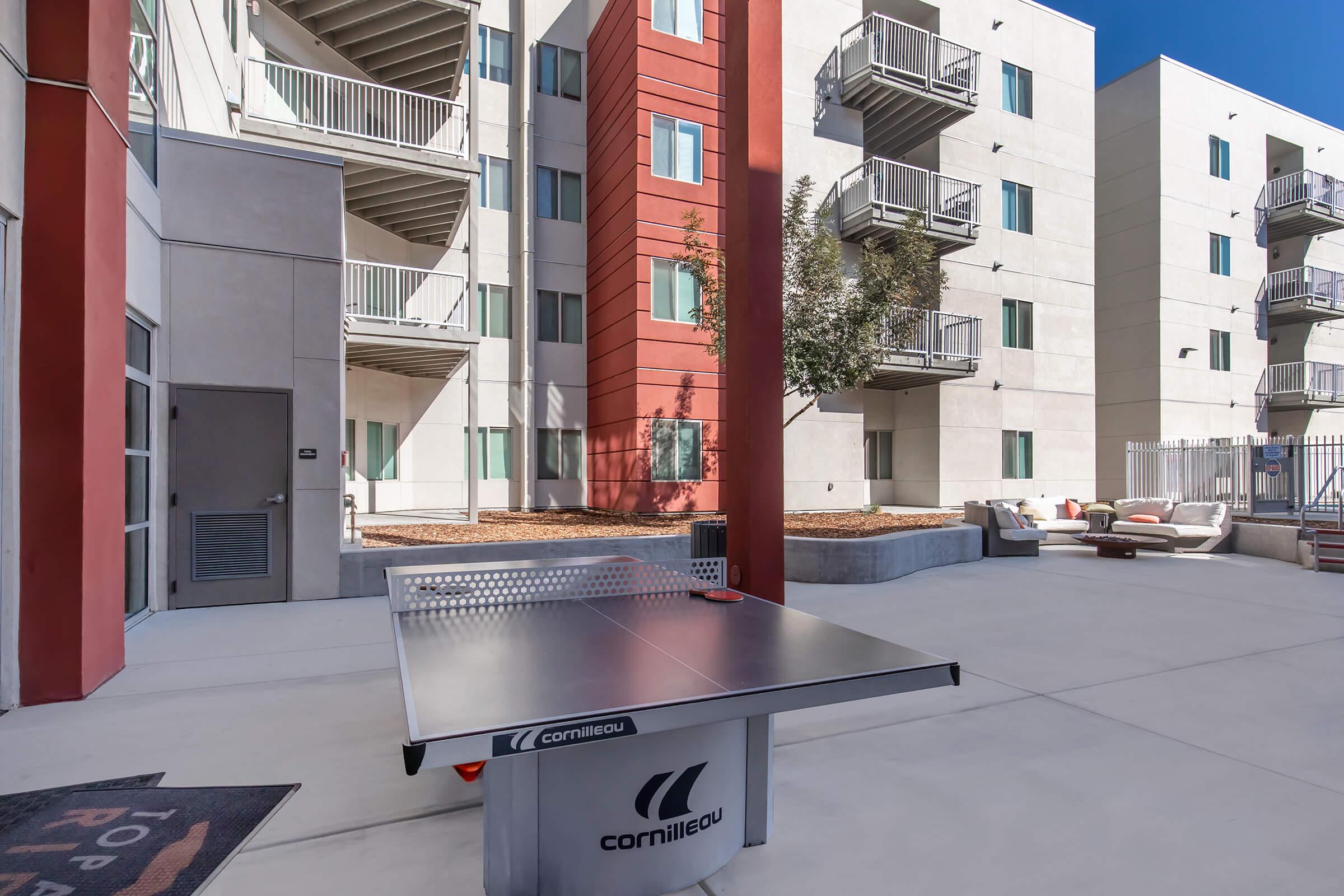 A table tennis setup in an outdoor communal area of an apartment complex. The background features modern buildings with balconies, and there are seating areas nearby. The space is well-lit, with clear blue skies overhead.