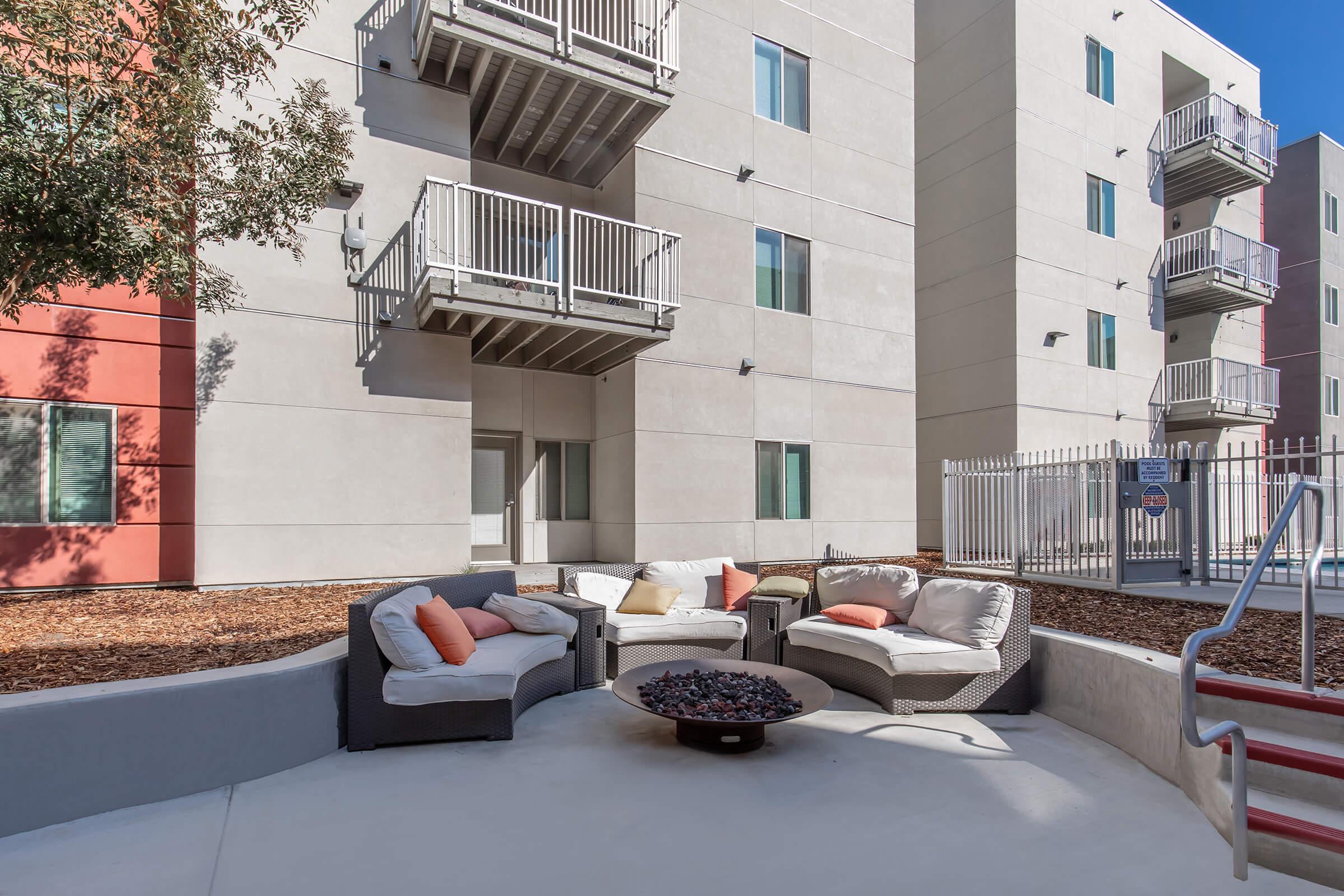 A modern outdoor seating area featuring comfortable couches with colorful throw pillows, surrounded by landscaped gravel. In the background, a contemporary multi-story building with balconies can be seen. A fire pit is centrally located on the patio, creating a cozy gathering space.