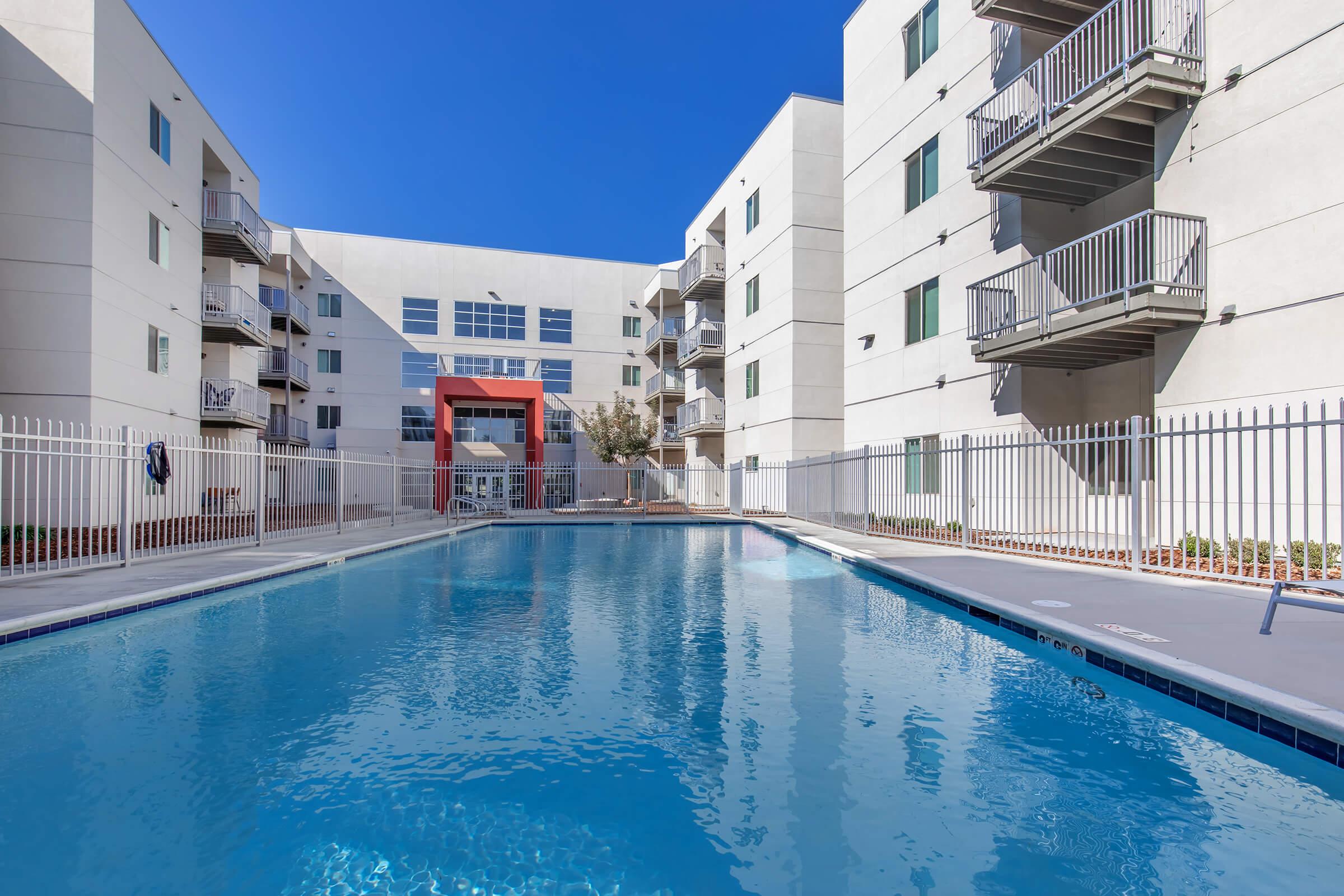 A clear blue swimming pool surrounded by a white fence, with modern apartment buildings in the background. The buildings feature balconies and large windows. The scene is set under a bright blue sky, creating a serene atmosphere for relaxation.