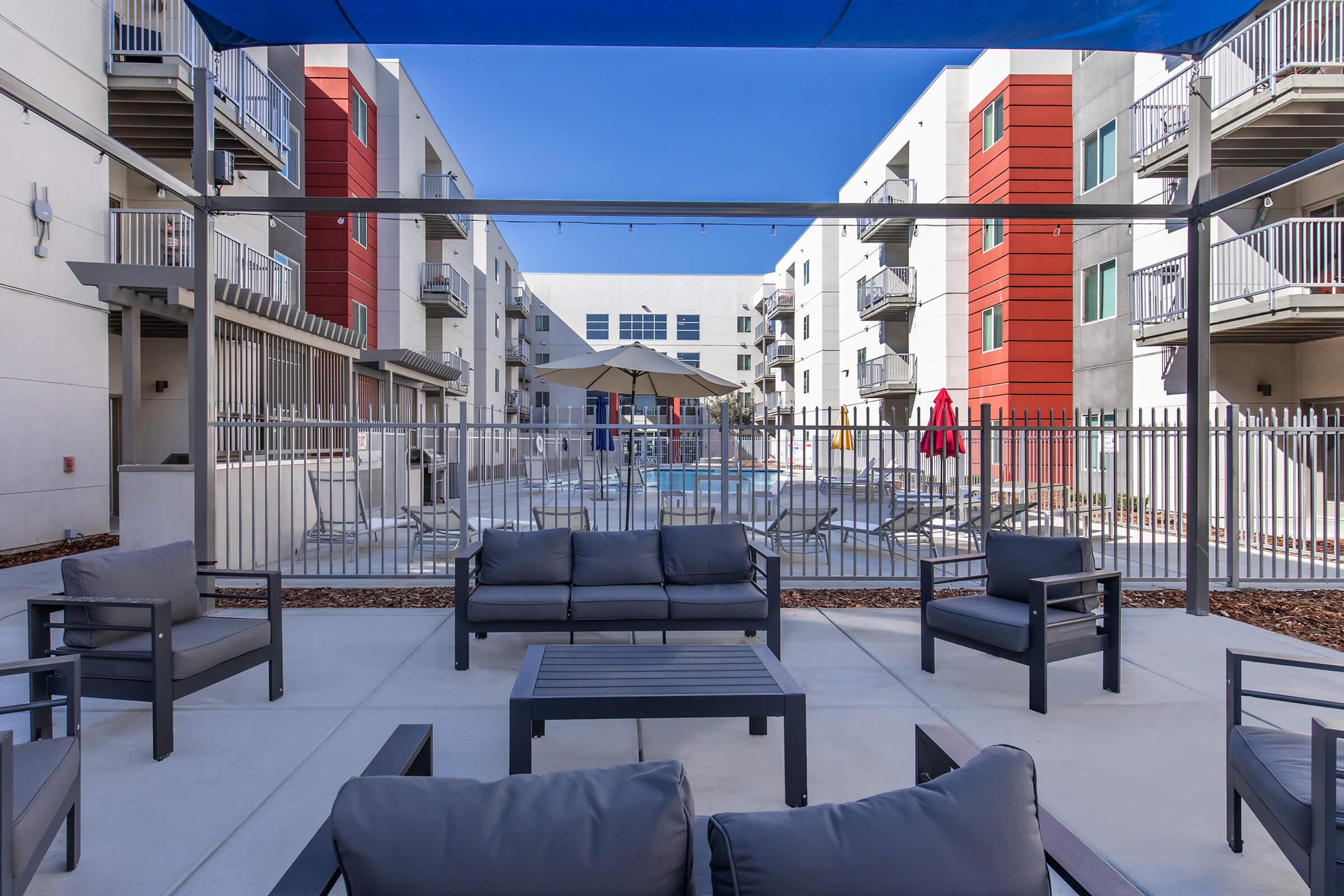 A modern outdoor lounge area featuring comfortable gray sofas and a coffee table, overlooking a swimming pool surrounded by a fenced area and shaded by a blue canopy. The background includes contemporary apartment buildings with balconies and a clear blue sky.