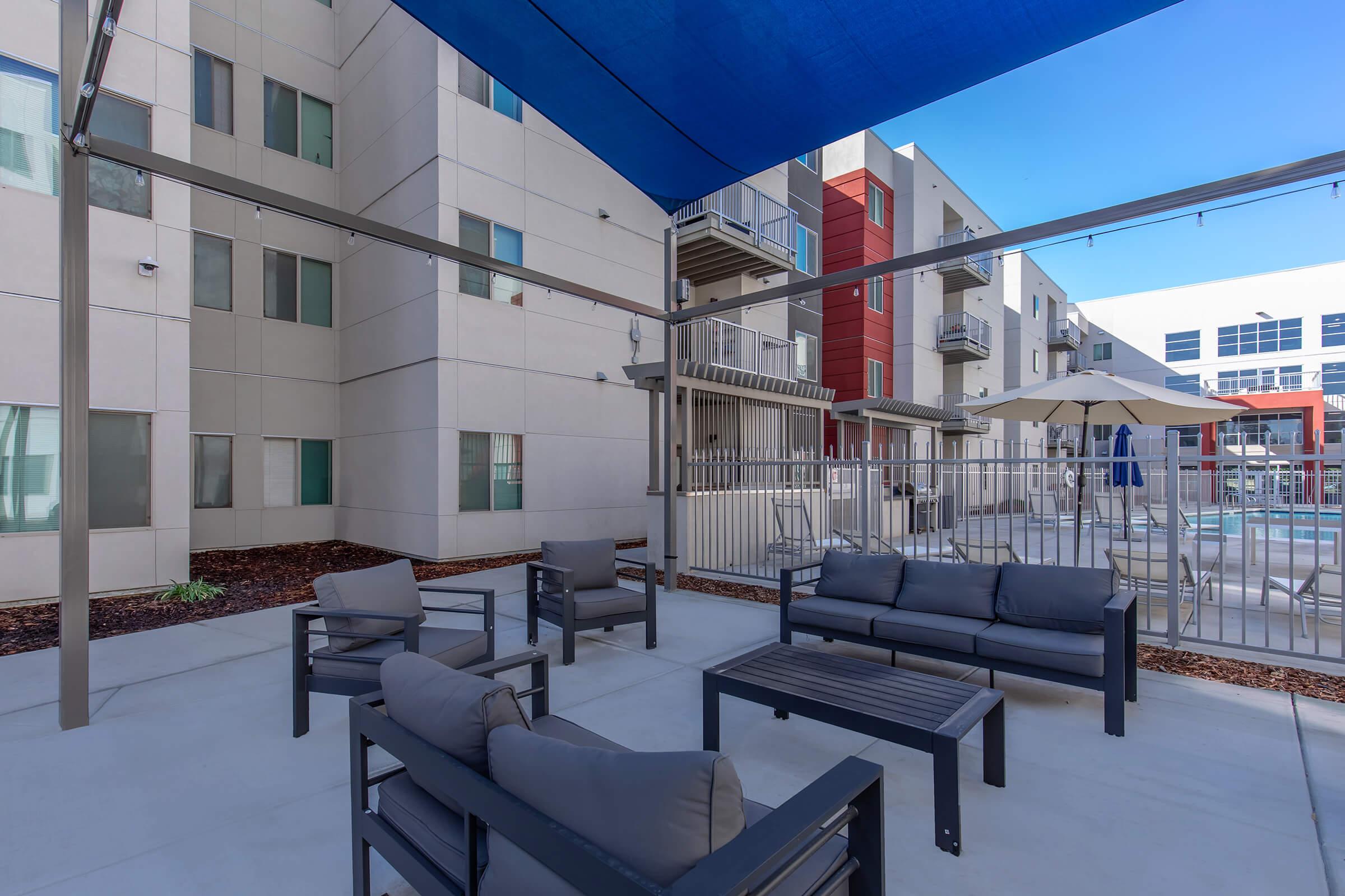 Outdoor seating area with gray sofas and black tables shaded by a blue canopy. In the background, a swimming pool is visible along with modern apartment buildings featuring balconies and large windows. The surroundings include landscaped elements and a fence around the pool area.