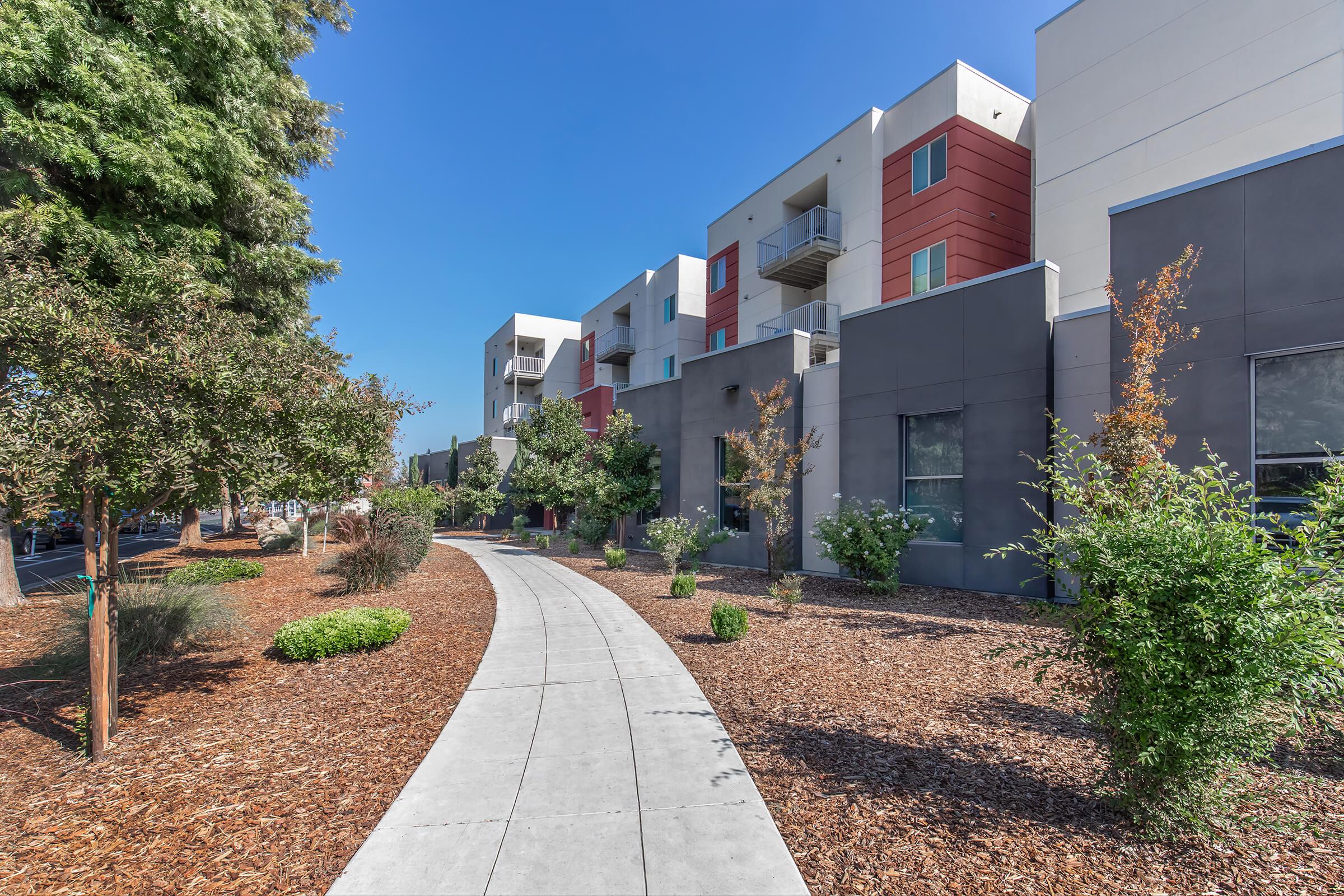 A sunny pathway lined with landscaped greenery leads to modern multi-story residential buildings with a mix of white, red, and gray façades. The scene conveys a welcoming and well-maintained urban environment.