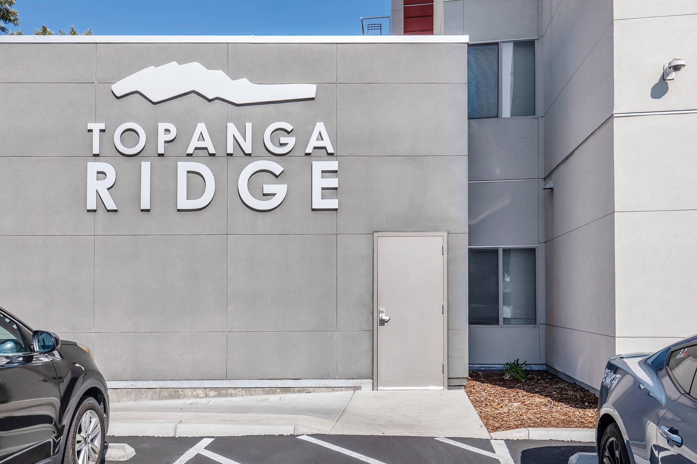 A modern building exterior featuring the sign "Topanga Ridge" mounted on a gray wall. The entrance has a simple door, and there are parking spaces in front of the building, with two parked cars visible. Sunlight illuminates the scene, creating a clean and inviting atmosphere.