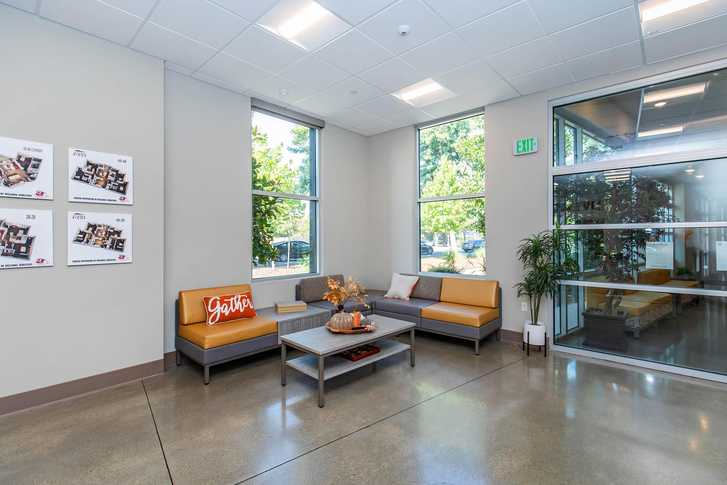 A modern lobby area featuring a comfortable seating arrangement with a gray and orange couch, a coffee table with decorative items, and large windows providing natural light. There are also informational boards on the wall and greenery visible outside, creating a welcoming atmosphere.