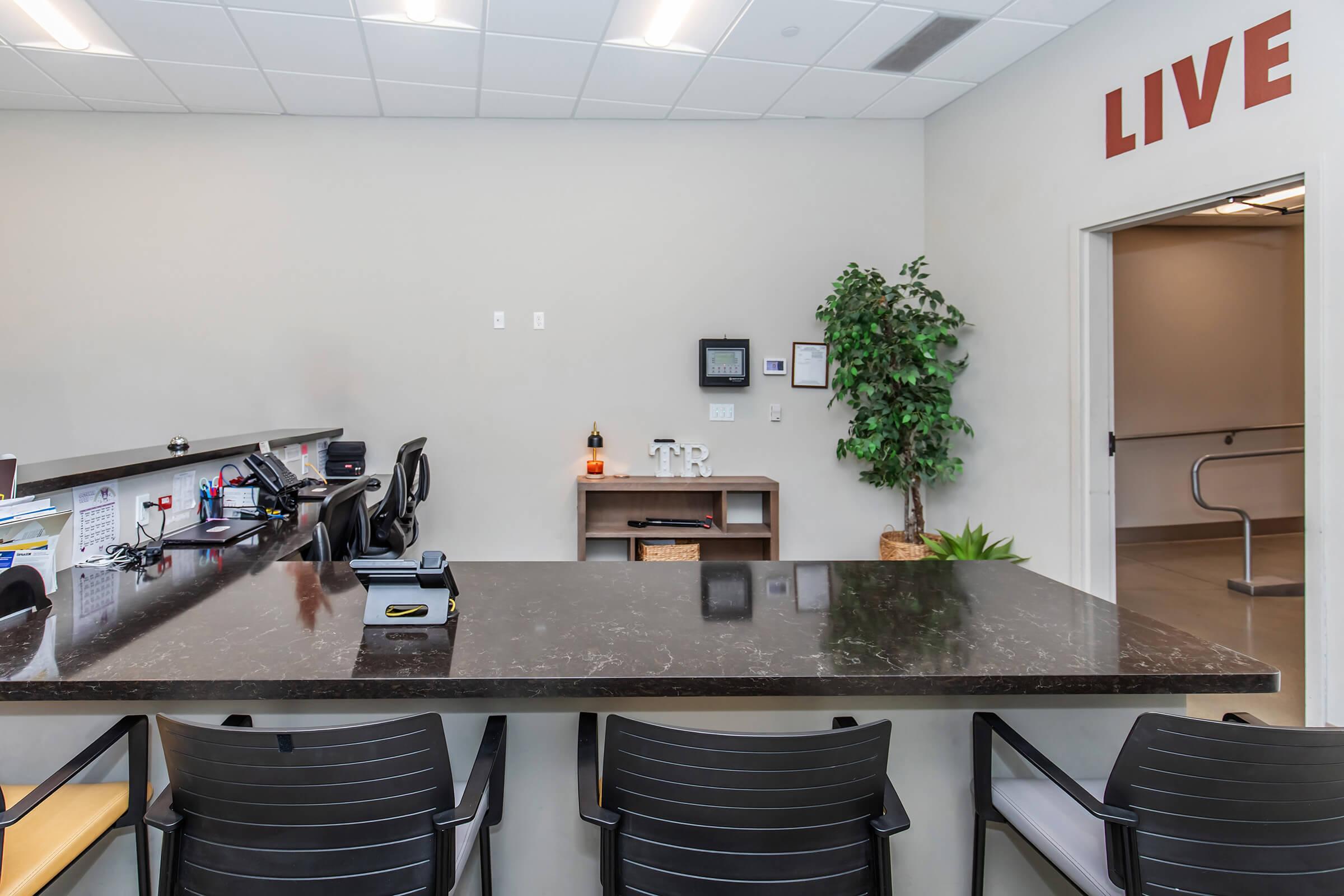 Reception area with a dark countertop, several black chairs, a potted plant, and a small shelf displaying decorative items. A wall features the word "LIVE" in large red letters. There are office supplies and a phone on the countertop, suggesting a professional or medical environment.