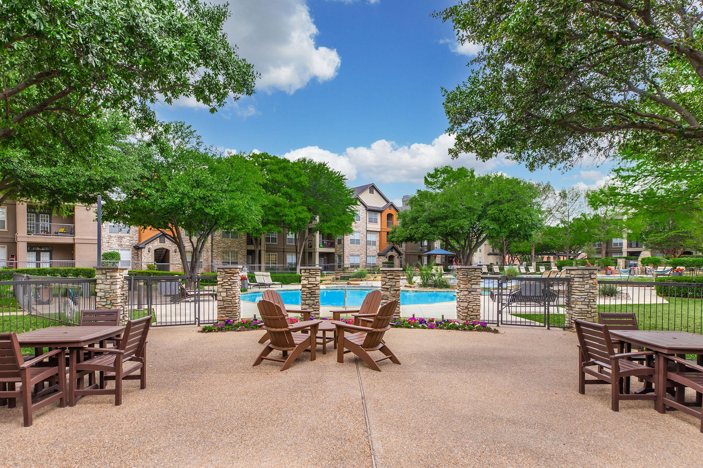 A landscaped pool area surrounded by green trees, featuring lounge chairs and outdoor tables. Apartments are visible in the background, with a clear blue sky and fluffy clouds above. Colorful flowers line the pathway, creating a welcoming atmosphere for residents and visitors.
