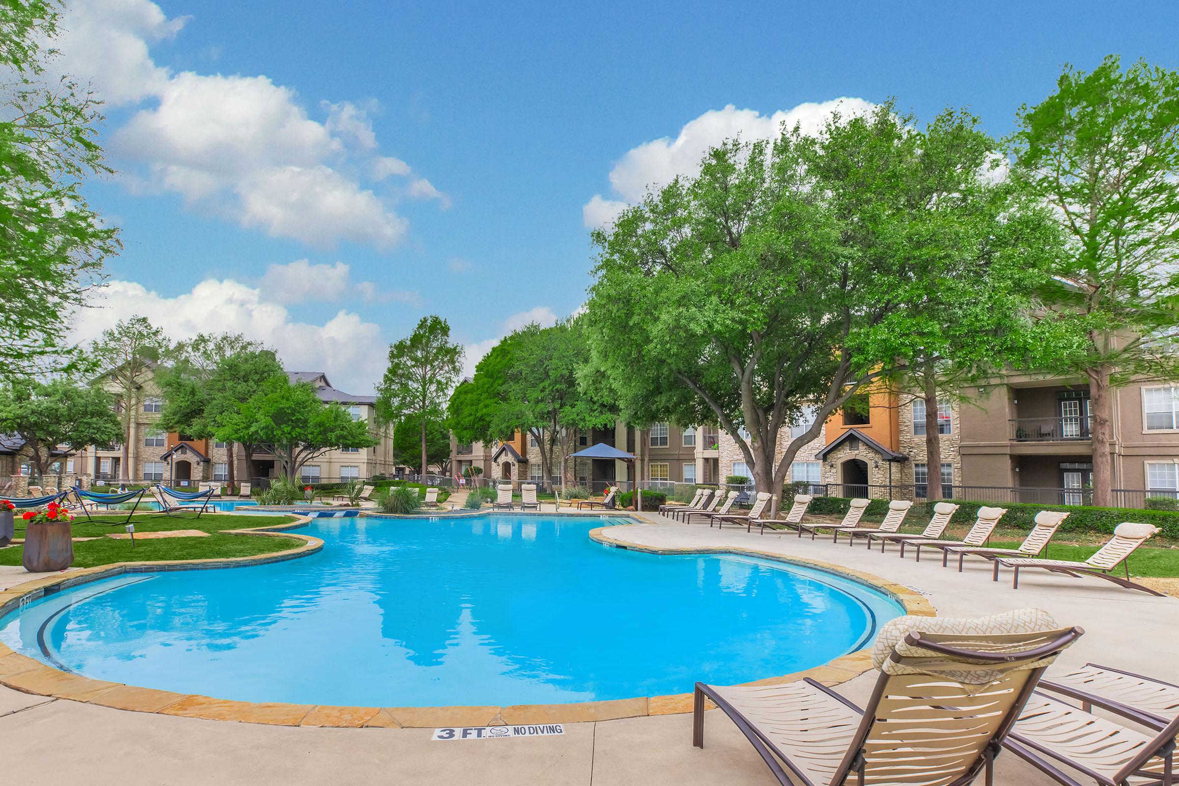 A serene outdoor swimming pool surrounded by lush greenery and lounge chairs. The pool is brightly lit under a clear blue sky with fluffy clouds, and residential buildings are visible in the background, creating a relaxing atmosphere.