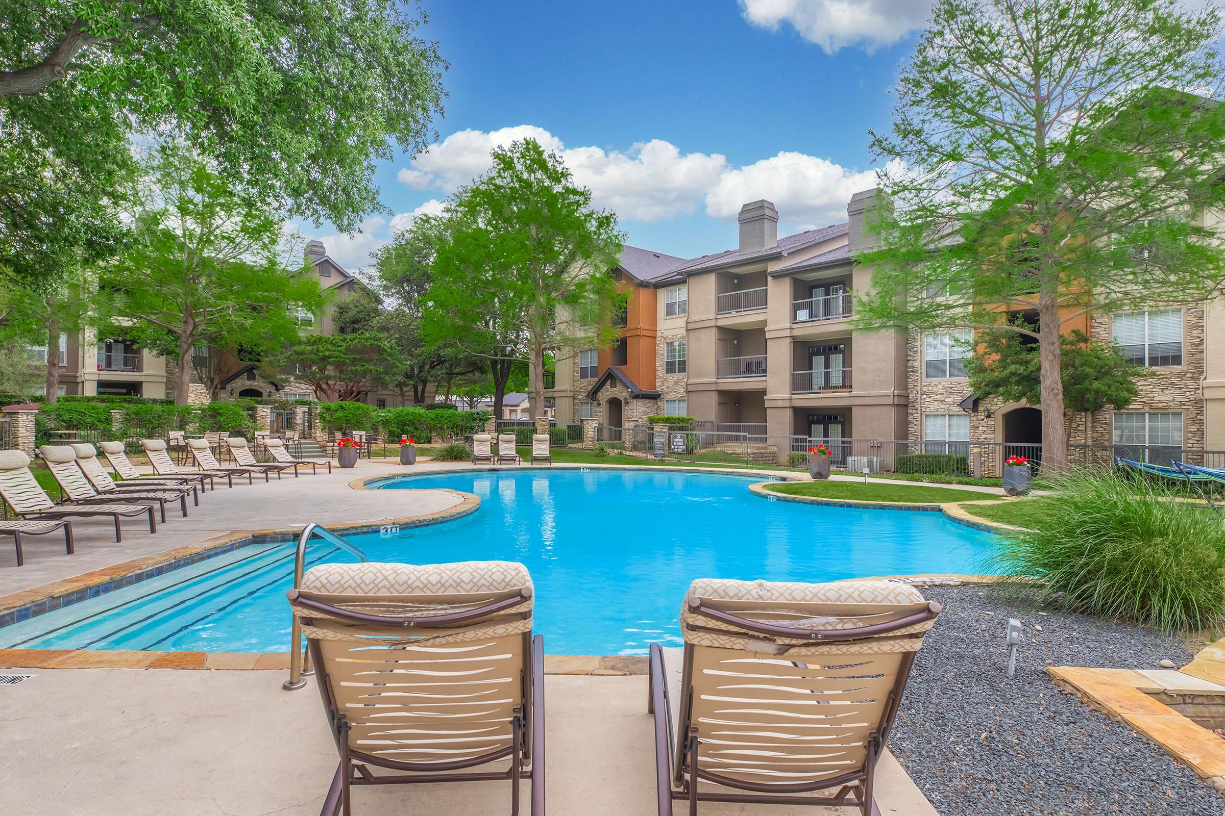 A serene apartment complex pool area featuring a bright blue swimming pool surrounded by landscaped greenery, lounge chairs, and trees. The sky is partly cloudy, and modern buildings are visible in the background, creating a relaxing and inviting atmosphere.