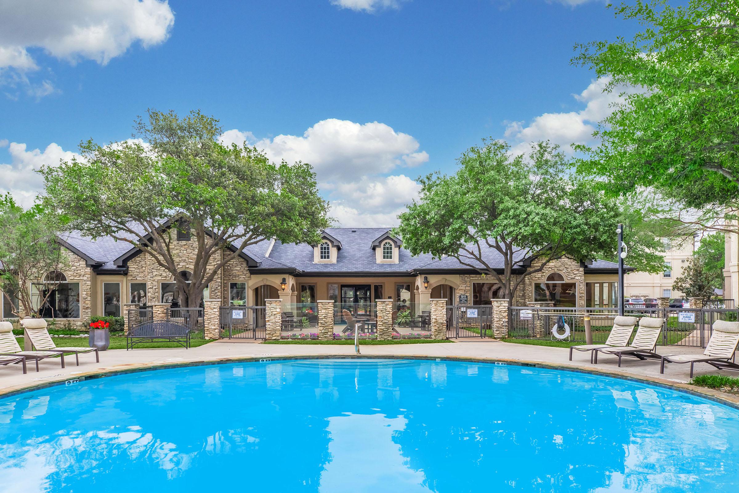A sunny pool area with lounge chairs surrounding a clear blue pool. In the background, a two-story building featuring stone accents and large windows is visible, framed by lush green trees and a bright blue sky with scattered clouds. Perfect for outdoor relaxation and leisure.