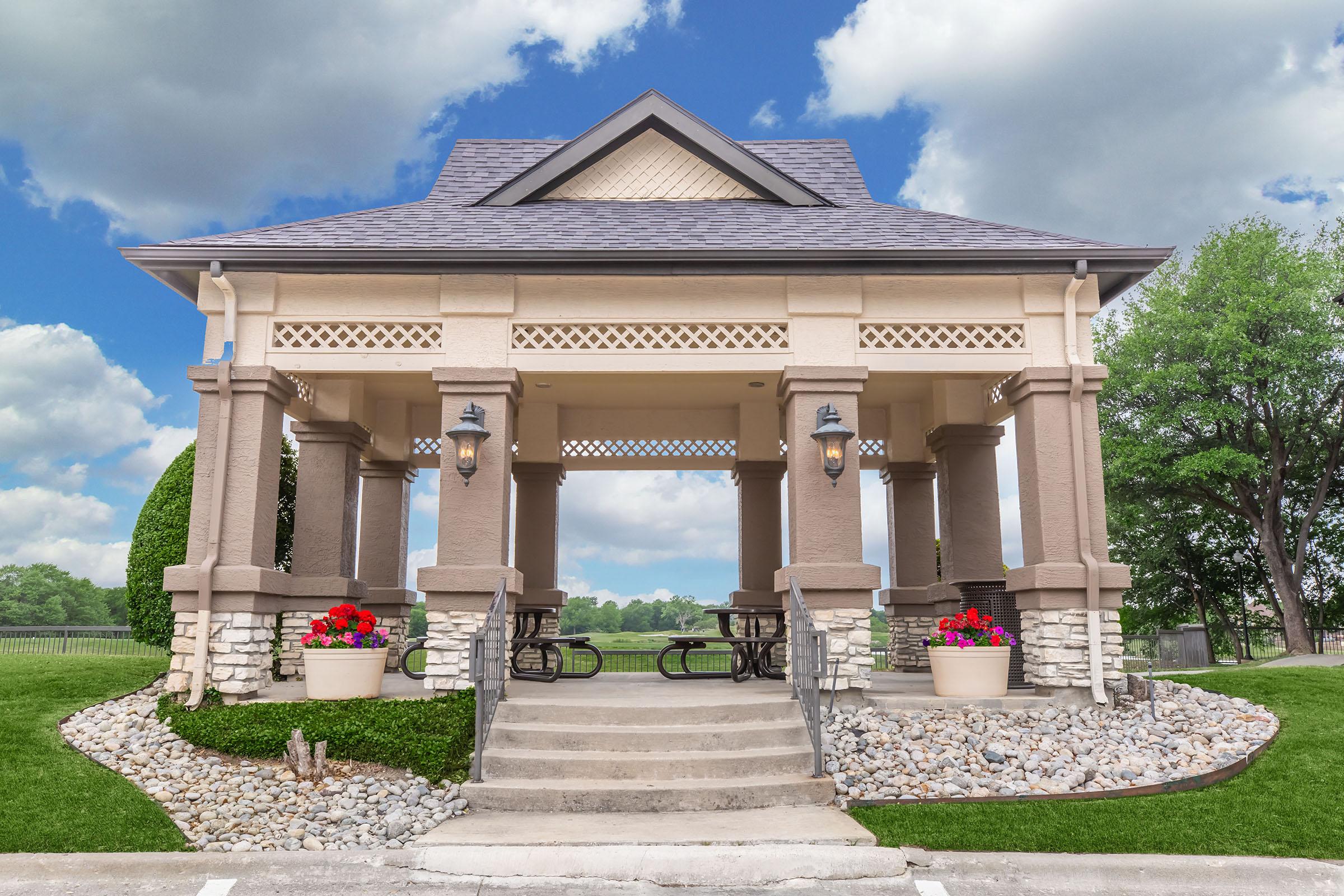 A charming gazebo with a peaked roof and decorative columns, surrounded by lush greenery. The entrance features steps leading up to a seating area with picnic tables. Flower pots with vibrant blooms are placed on either side, adding color to the scene, under a partly cloudy sky.