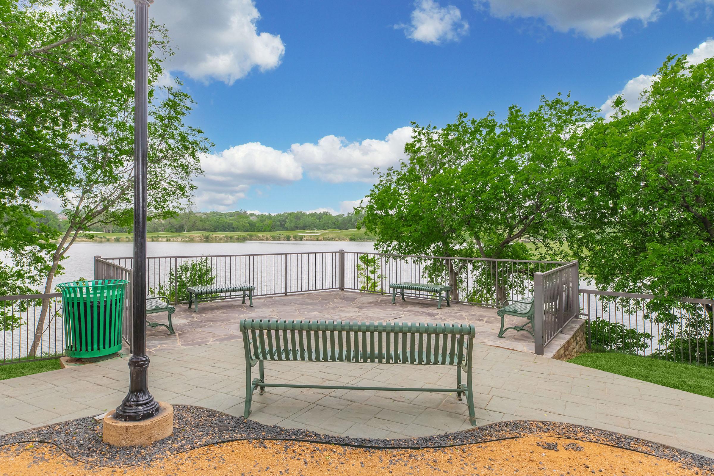 A scenic view from a park overlooking a river, with green trees framing the area. There are several benches for sitting, a trash bin nearby, and a pathway made of stone and gravel. The sky is partly cloudy, creating a serene outdoor atmosphere.