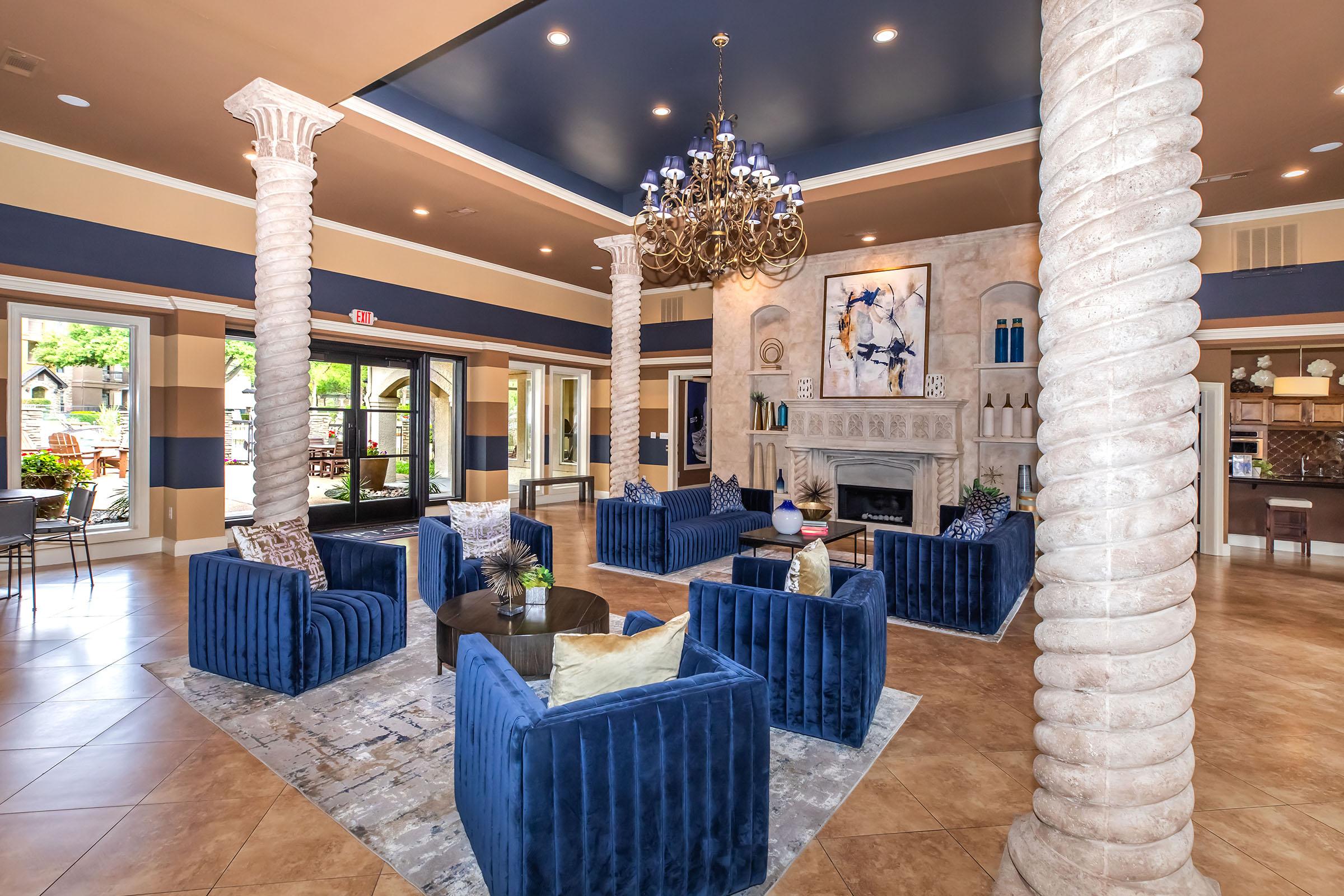 A modern, elegant lobby featuring tall columns, a chandelier, and blue velvet furniture. The decor includes a large abstract painting and a cozy fireplace. Natural light streams in through large windows, creating a welcoming atmosphere. Stylish accents and warm tones enhance the inviting setting.