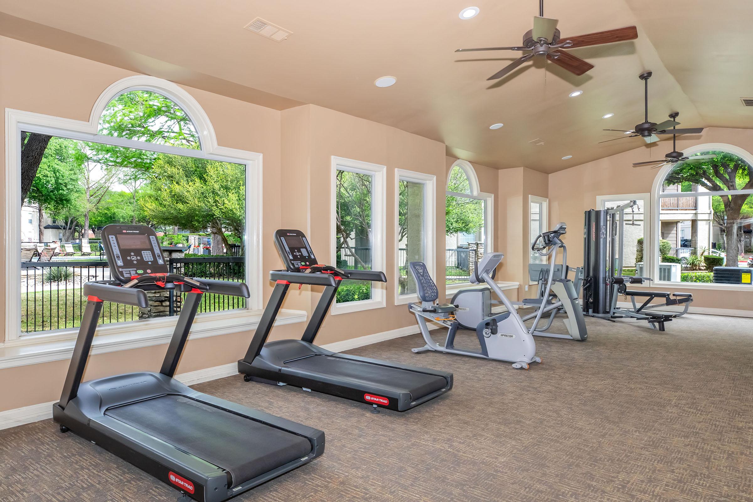 A well-lit gym featuring two treadmills and a versatile workout machine, positioned near large windows that offer a view of green trees outside. The room has a neutral color scheme with ceiling fans, providing a spacious and inviting exercise environment.