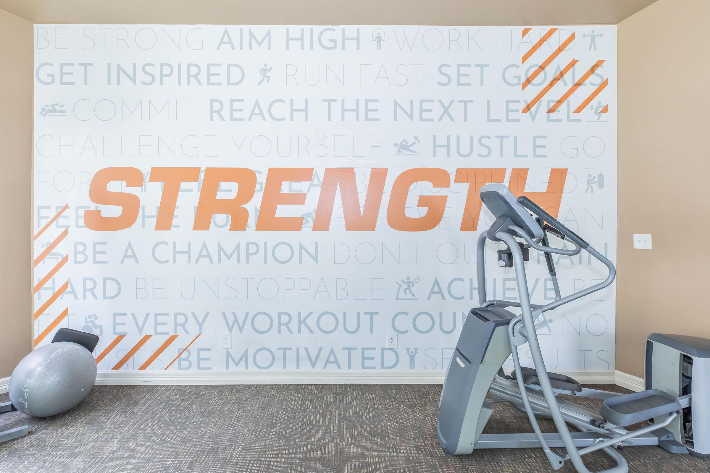 A fitness studio wall featuring motivational phrases and the word "STRENGTH" prominently displayed in large orange letters. The background includes various fitness-related words and icons. A treadmill and a stability ball are visible in the foreground, emphasizing an active workout environment.