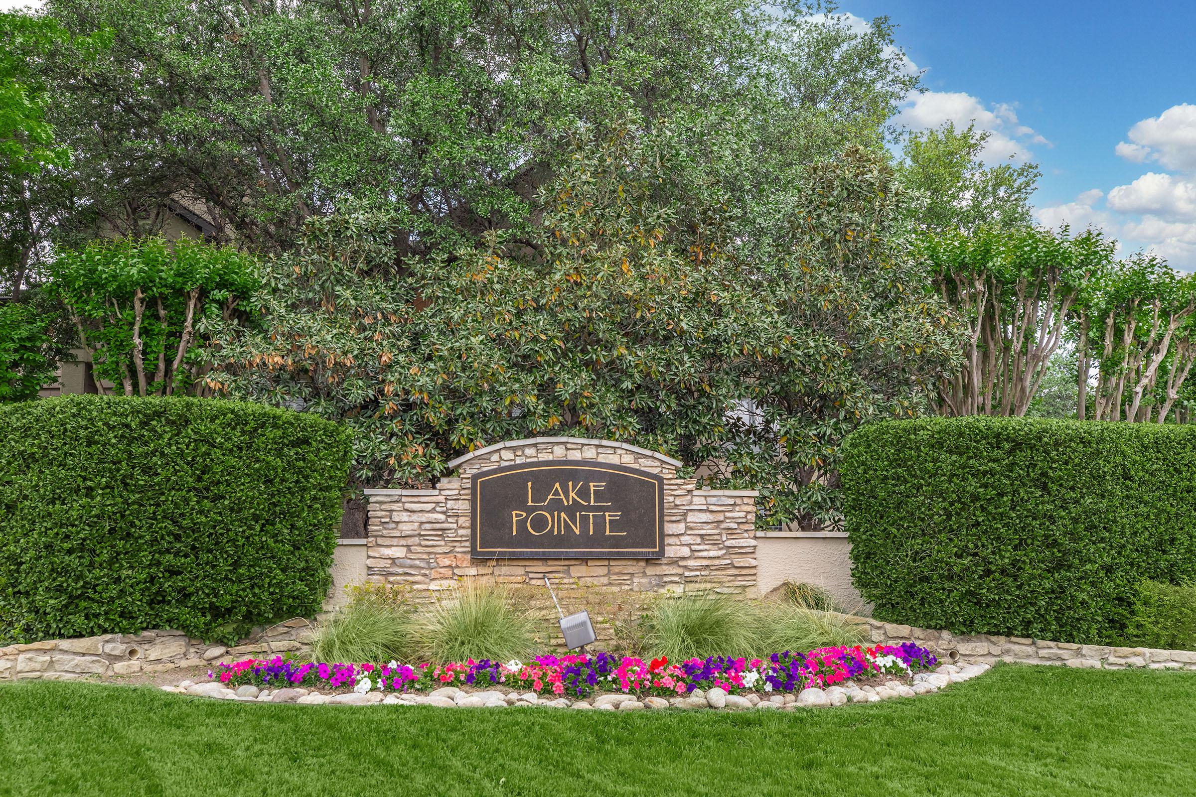 A welcoming entrance to Lake Pointe featuring a decorative stone sign surrounded by lush greenery and colorful flower beds. The sign is prominently displayed amidst well-trimmed hedges, creating an inviting atmosphere. The sky is partly cloudy, adding to the serene setting.
