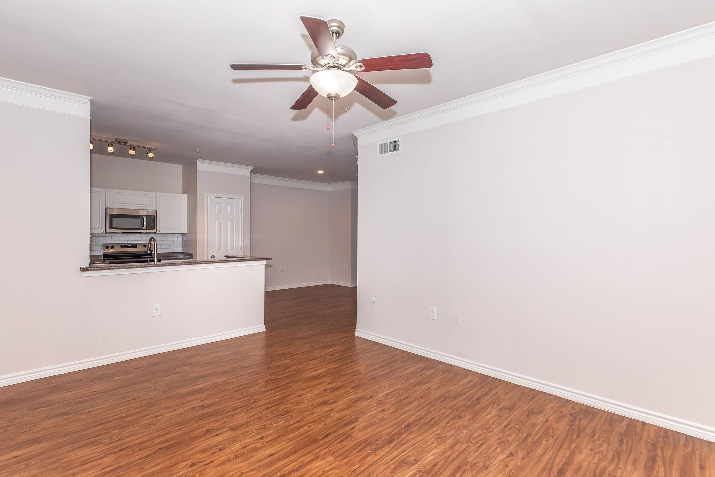 Spacious room with hardwood flooring, a ceiling fan, and neutral-colored walls. An open kitchen area is visible in the background, featuring stainless steel appliances. The layout suggests a modern, inviting living space with plenty of natural light and room for furniture arrangement.