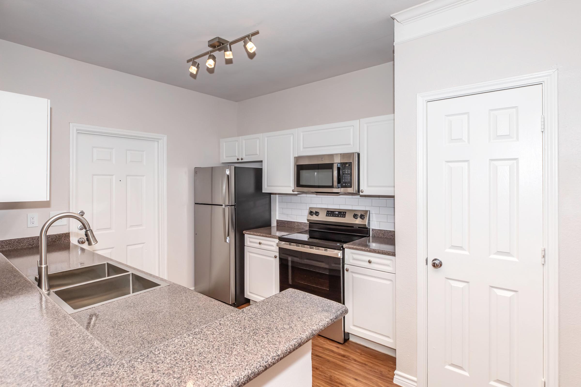 Modern kitchen featuring stainless steel appliances, including a refrigerator, microwave, and oven. The countertops are gray granite, and there is a double sink. White cabinets line the walls, and a door provides access to another room. The space has warm wood flooring and is well-lit with recessed lighting.