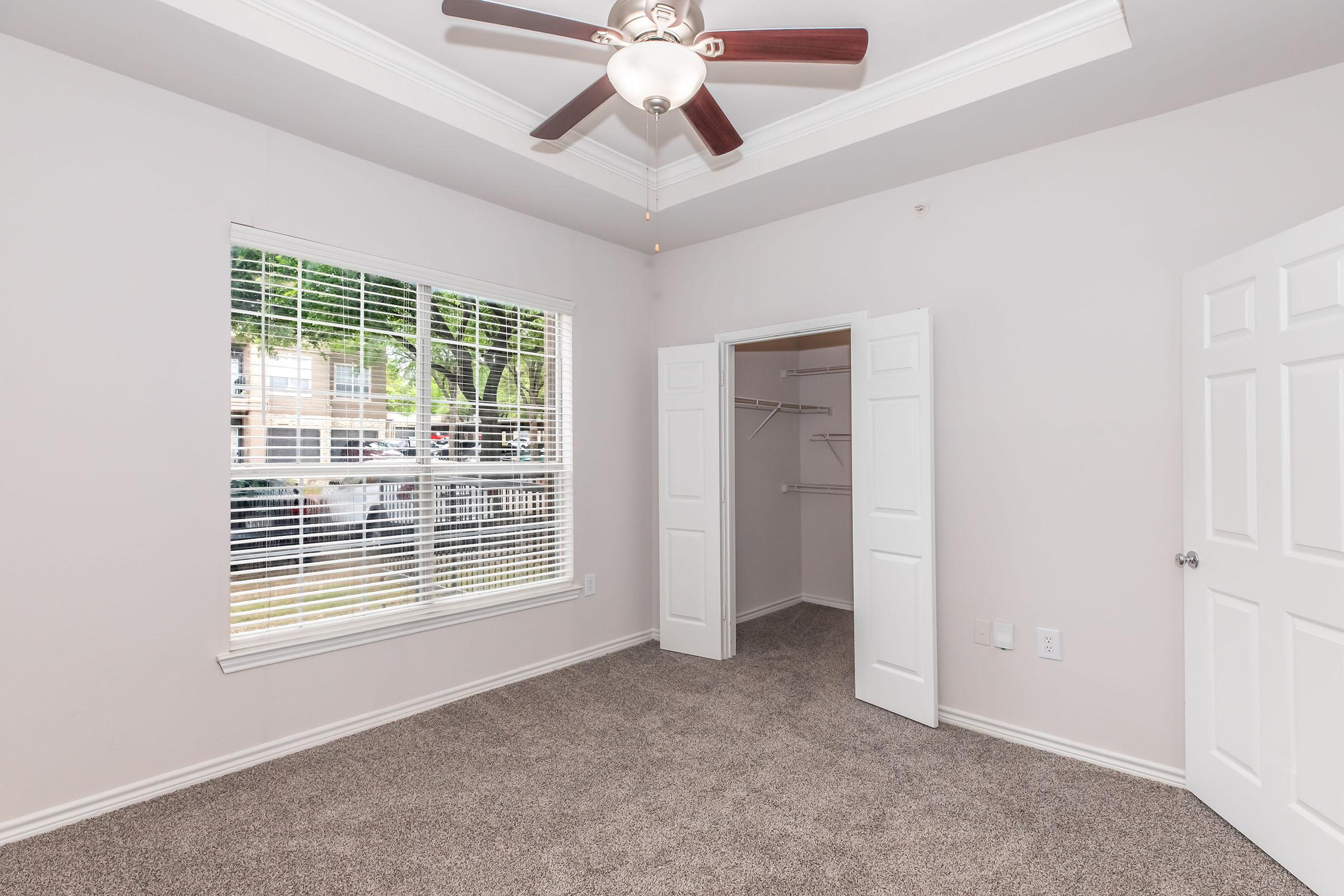 A spacious, light-filled room featuring a ceiling fan, beige walls, and plush carpeting. The room includes a window with blinds, allowing natural light to enter. There is a closet with double doors and shelving, providing storage space, and the view outside shows greenery and parked vehicles.