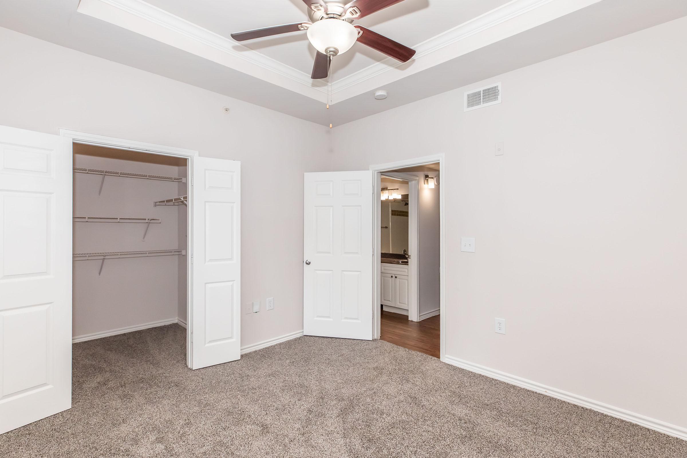 A spacious room featuring light-colored walls and carpet, with a ceiling fan. Two white double doors open to a closet on one side, and another door leads to a bathroom. The room has a modern design with natural light, creating a welcoming atmosphere.