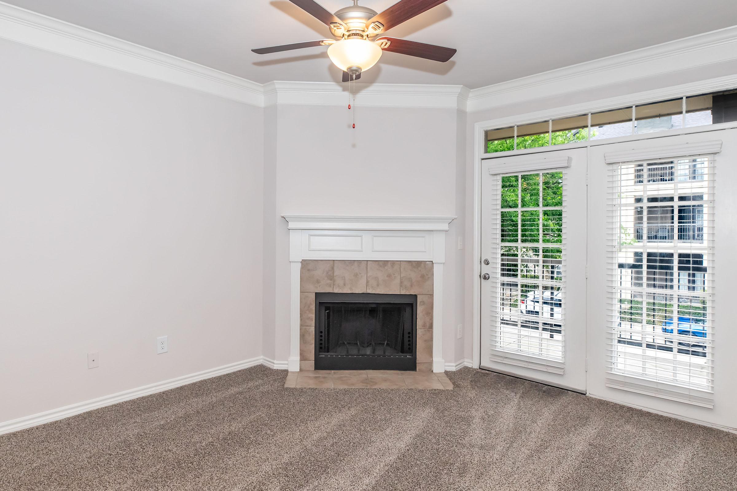 A cozy living room featuring a light-colored wall, a ceiling fan, and a fireplace with a beige surround. The room has plush carpet and sliding glass doors that lead to a balcony or patio area with a view of greenery. Natural light flows in through the windows, creating a welcoming atmosphere.