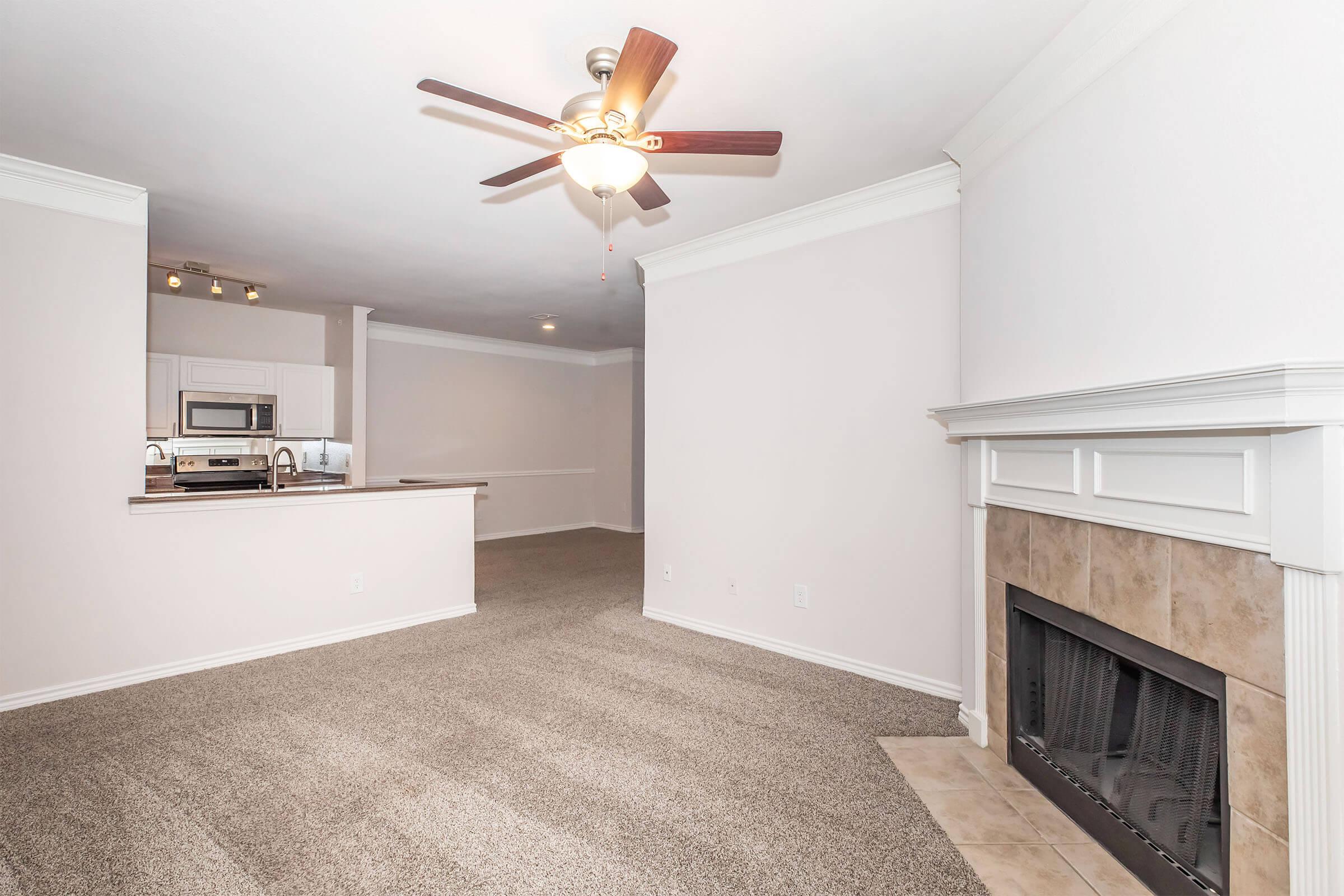 Spacious living area with plush carpeting, a ceiling fan, and a cozy fireplace. The room features light-colored walls and an open layout that connects to a kitchen area with modern appliances, offering a bright and inviting atmosphere.