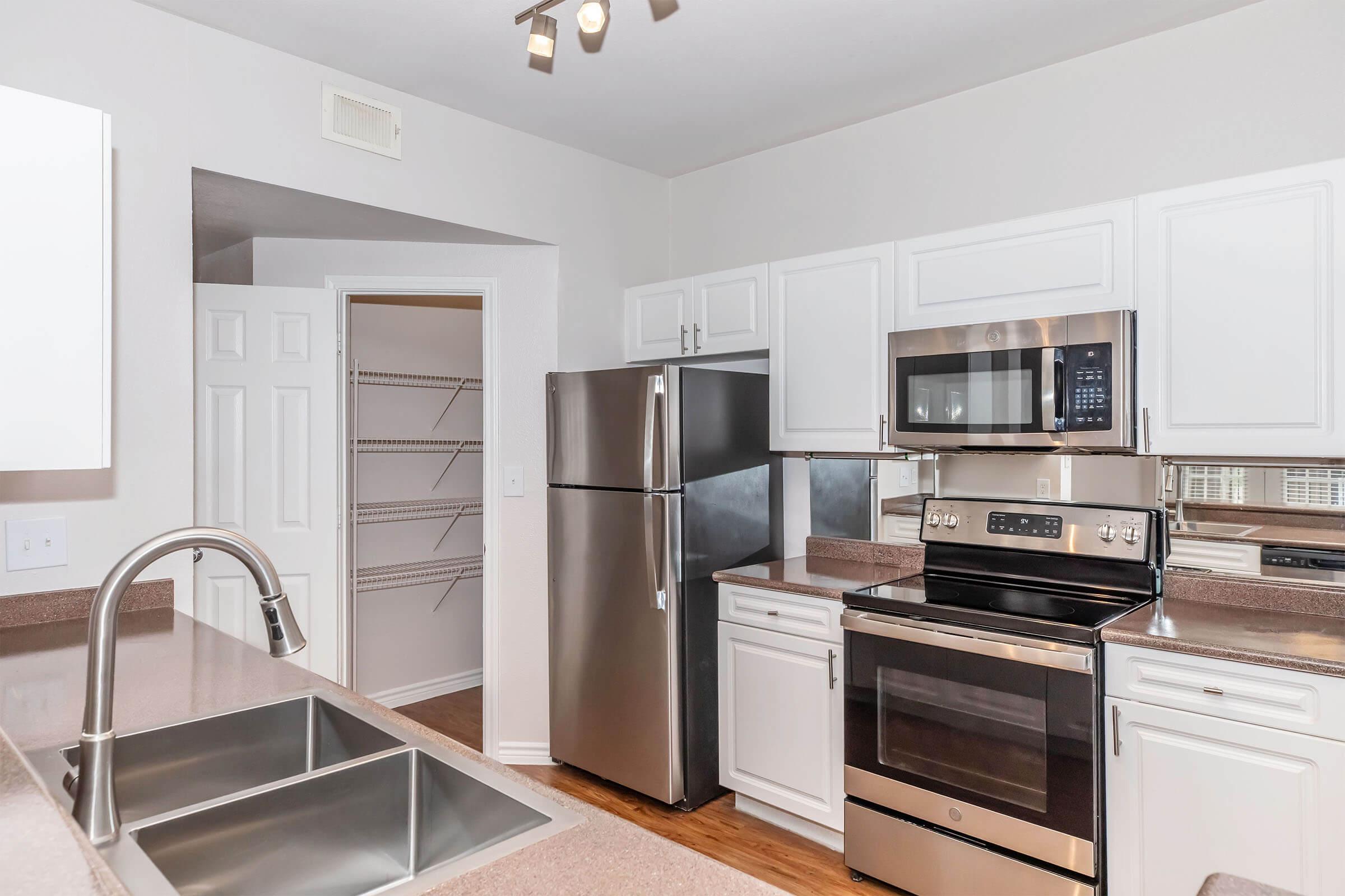 Modern kitchen featuring stainless steel appliances, including a refrigerator, microwave, and oven. White cabinetry and light countertops create a bright atmosphere. A pantry can be seen through an open door, and the sink area offers a double basin. The floor is finished with warm wood tones.