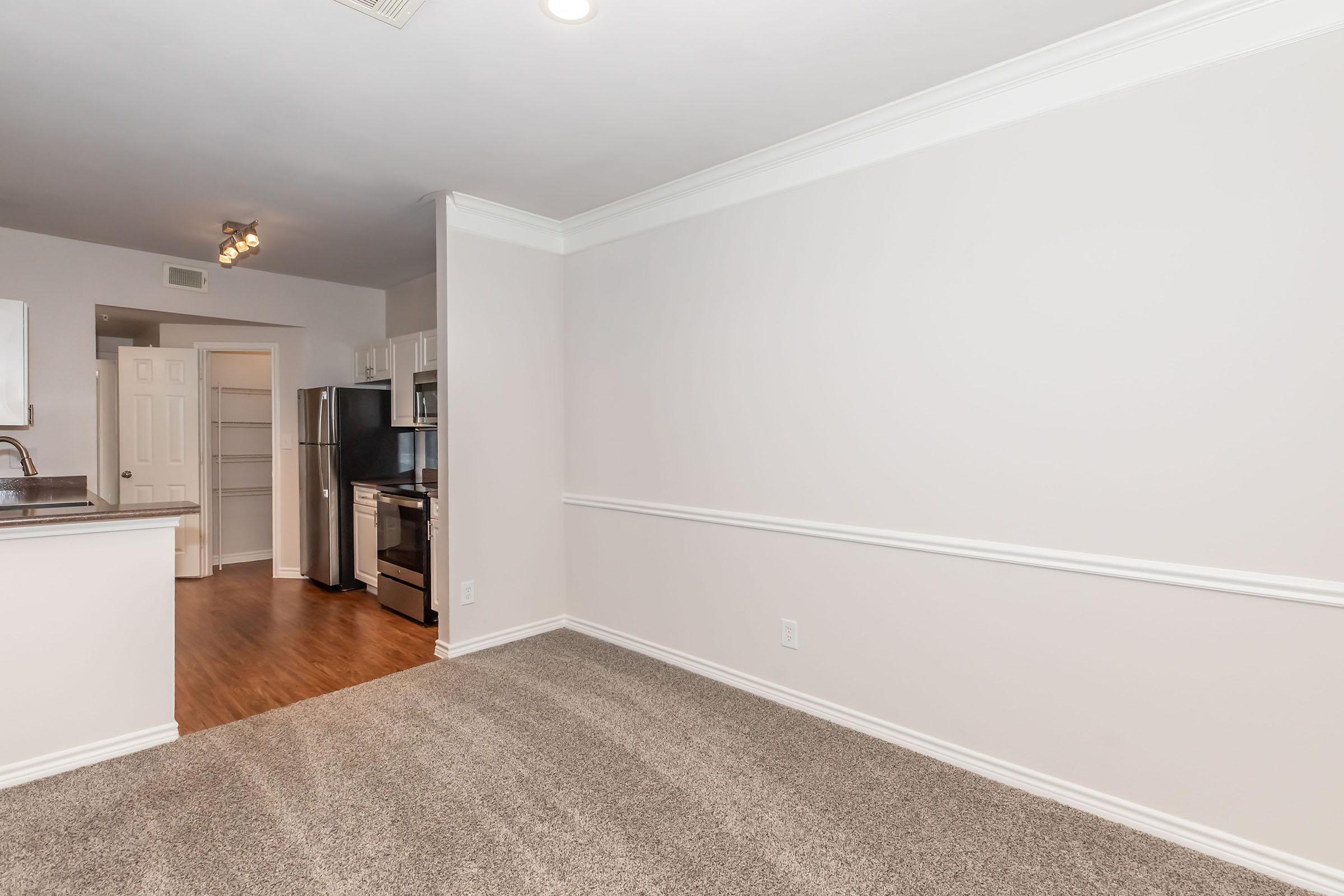 A spacious room with light-colored walls and carpet flooring, featuring a doorway leading to a kitchen area. The kitchen is equipped with modern stainless steel appliances and offers an open layout. Natural light from overhead fixtures illuminates the space, creating a welcoming atmosphere.