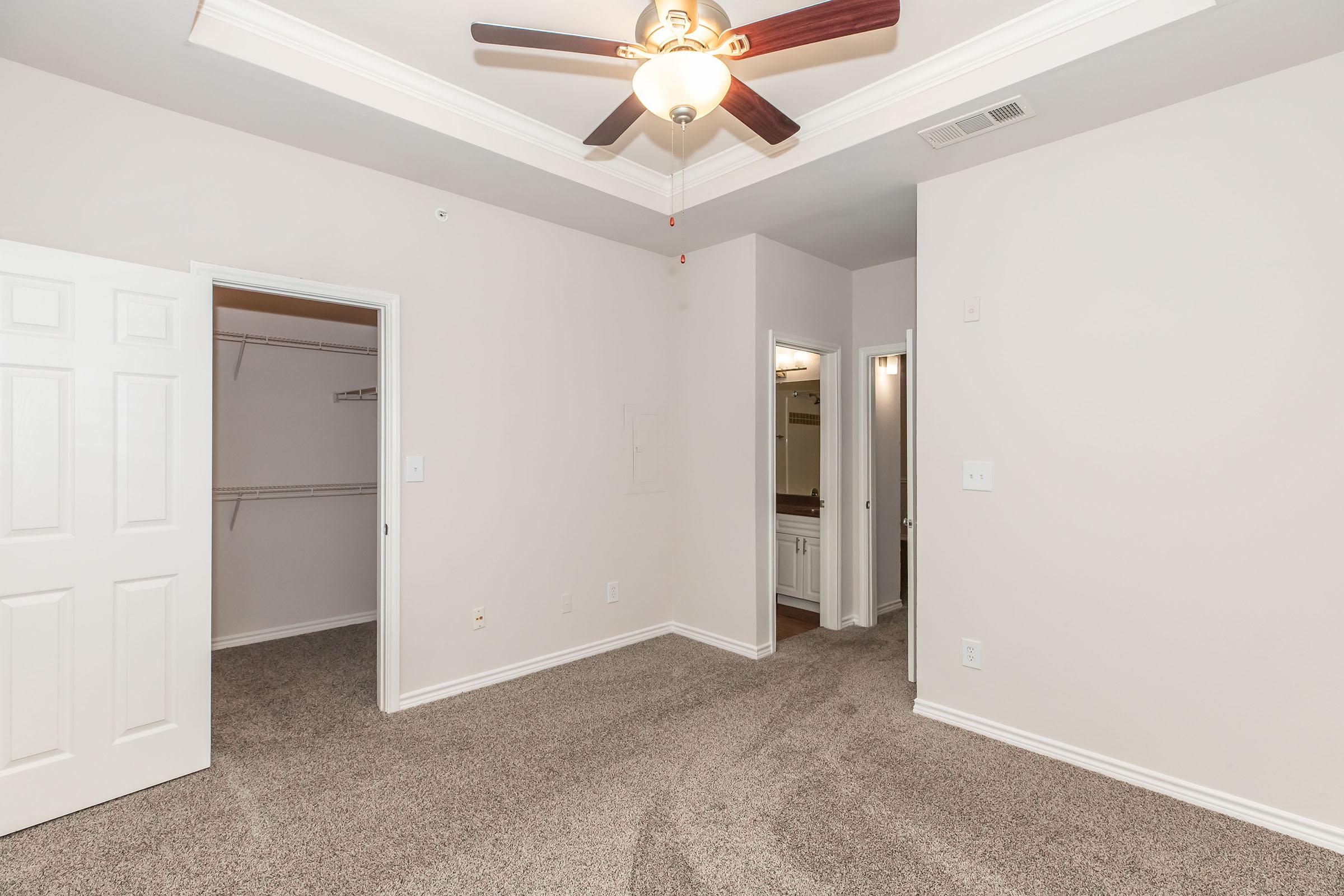 A spacious bedroom featuring light beige walls and carpet. The room has a ceiling fan with light, a closet with double doors, and two doorways leading to other areas. Natural light comes from the openings, creating a bright and inviting atmosphere.