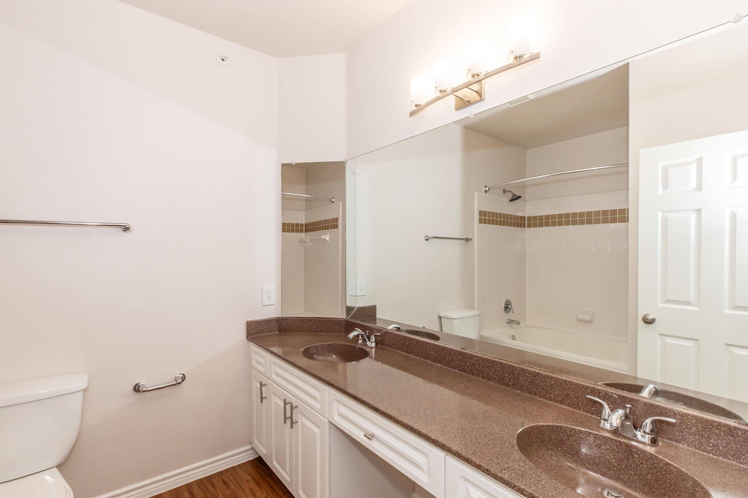 A clean, modern bathroom featuring a large mirror above a dual vanity with granite countertops, two sinks, and chrome faucets. There is a white toilet and a shower area with glass doors. The walls are painted a light color, and the flooring is hardwood.