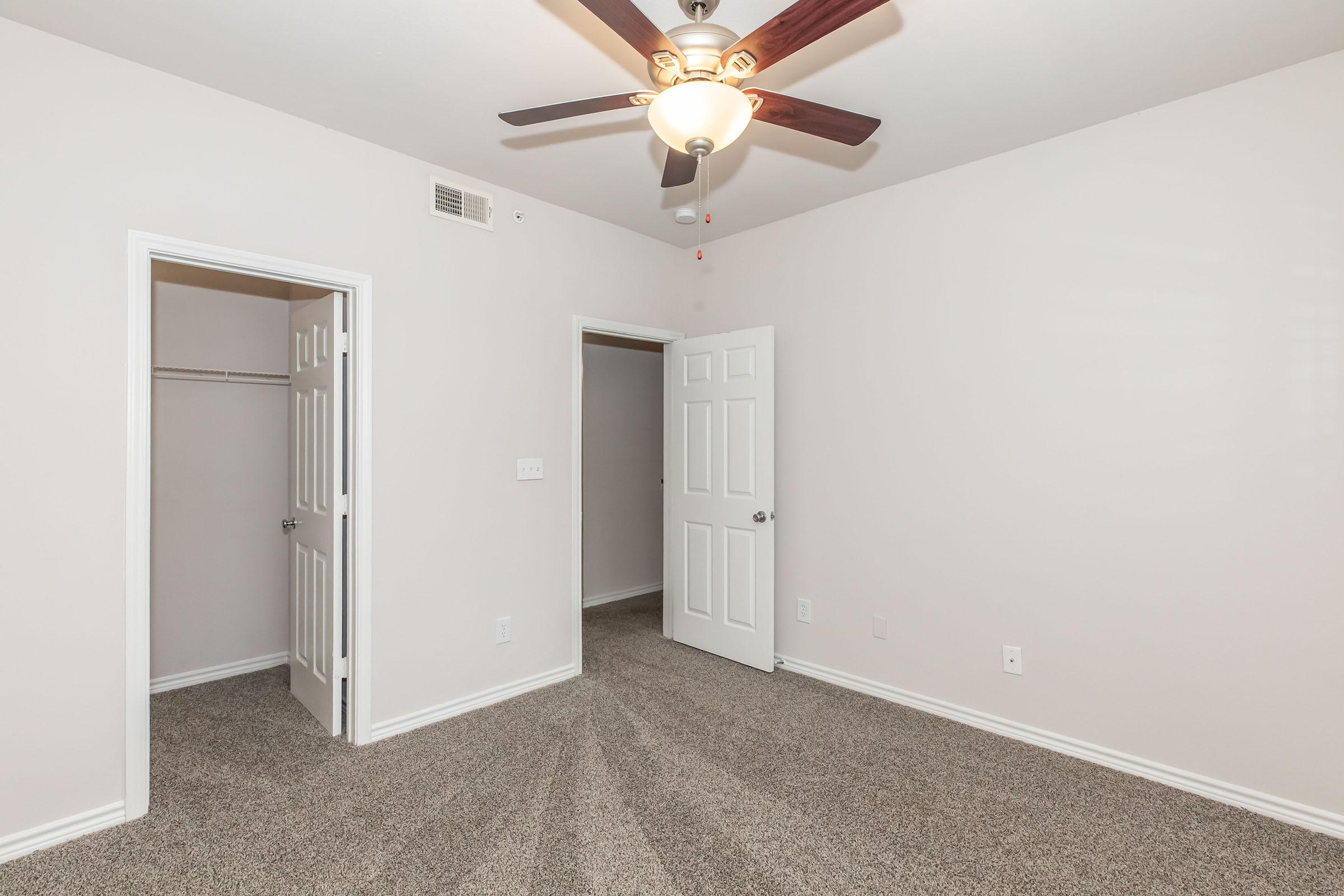 A clean, light-colored bedroom featuring a ceiling fan, carpeted flooring, and two white doors leading to closets. The walls are painted in a soft shade, creating a warm and inviting atmosphere. The room is spacious and well-lit, with natural light coming from a nearby window.
