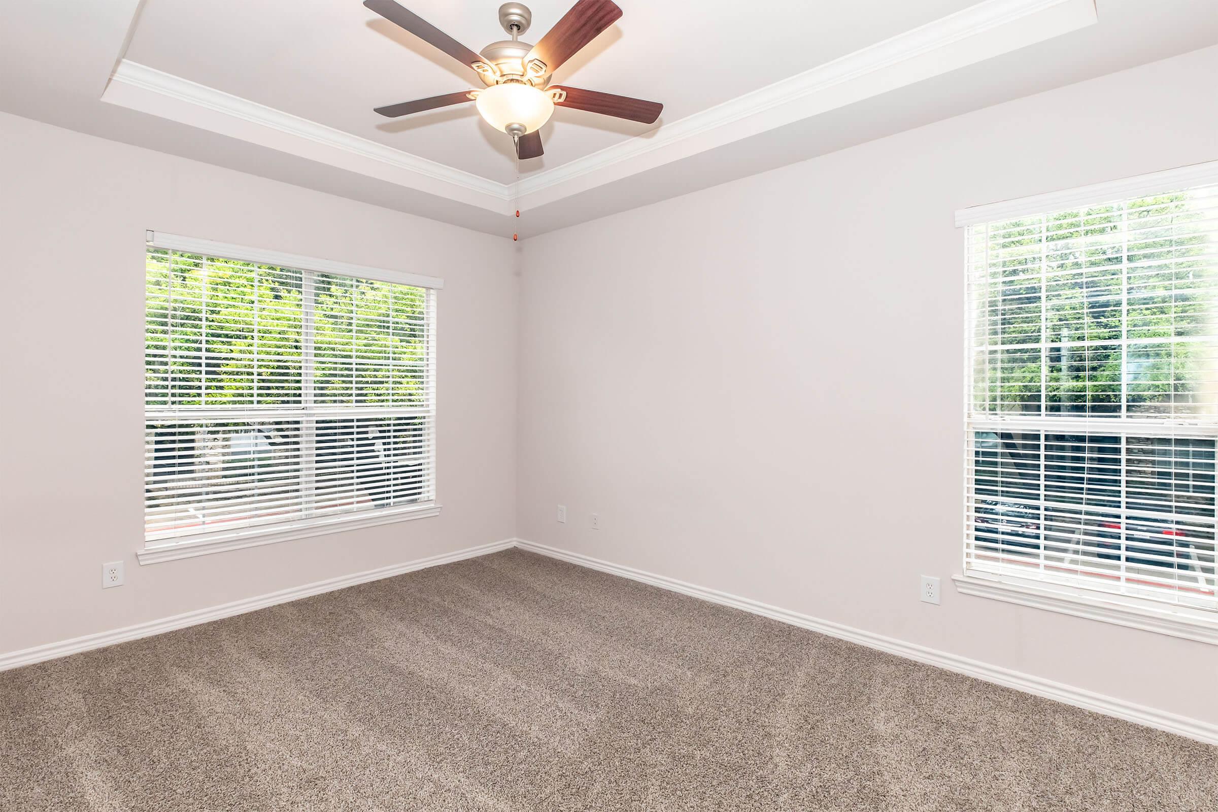 A bright, empty room featuring light-colored walls and plush carpet. There are two windows adorned with white blinds, allowing natural light to fill the space. A ceiling fan with elegant light fixture is mounted in the center, adding a modern touch to the otherwise minimalist design.