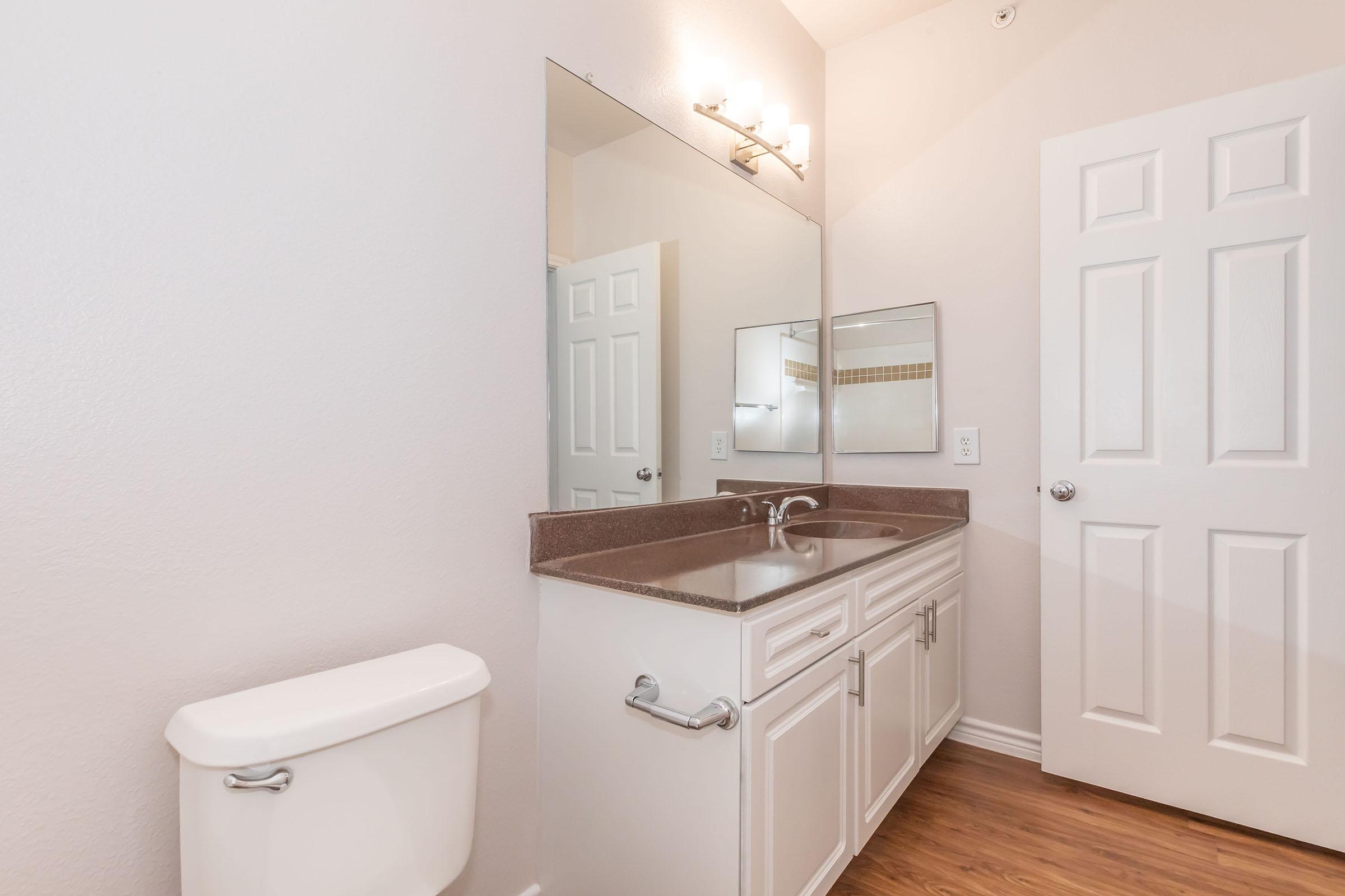 A clean and modern bathroom featuring a white vanity with a dark countertop, a large mirror above the sink, a toilet, and light-colored walls. The flooring is wooden, and there are two doors, one leading to the restroom area and another possibly to a storage or closet space.