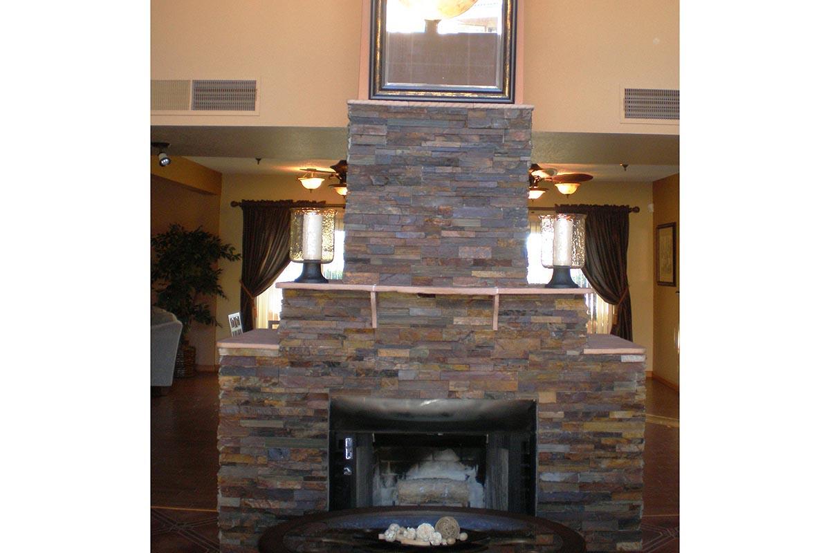 A stone fireplace with a dark wooden mantel, centered in a living room. Above the fireplace hangs a framed picture, and two lamps are positioned on either side of the mantel. The room features warm-colored walls, large windows with curtains, and a glimpse of furniture in the background.