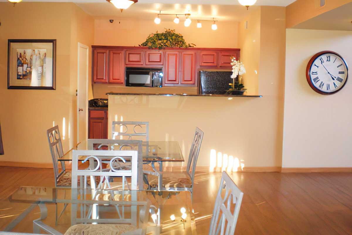 A modern kitchen with red cabinetry and a black countertop, featuring a clock on the wall and a framed artwork. There's a dining area with a glass table and stylish metal chairs, set in a bright, warmly lit room with wooden flooring. Soft, natural light filters through the space.