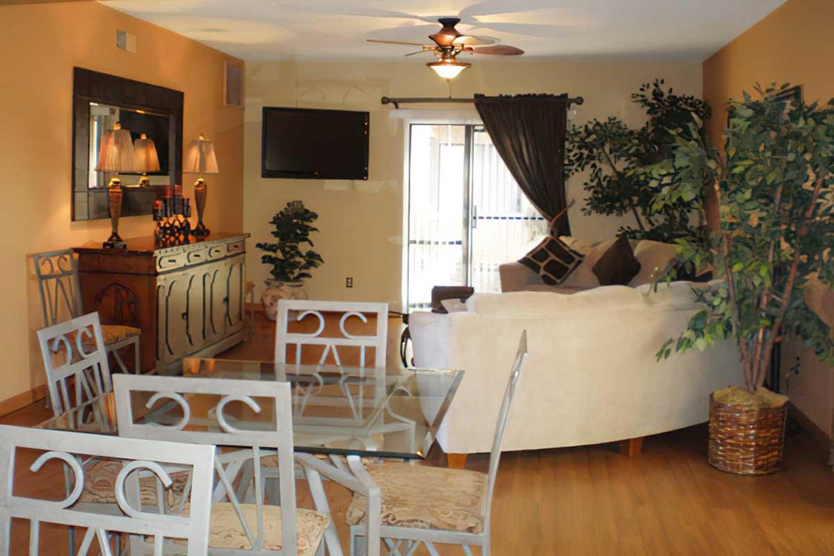 A cozy living space featuring a light-colored sofa, a glass dining table with decorative chairs, and a wooden sideboard. The room includes plants, a wall-mounted TV, and large windows with curtains, creating a bright and inviting atmosphere.