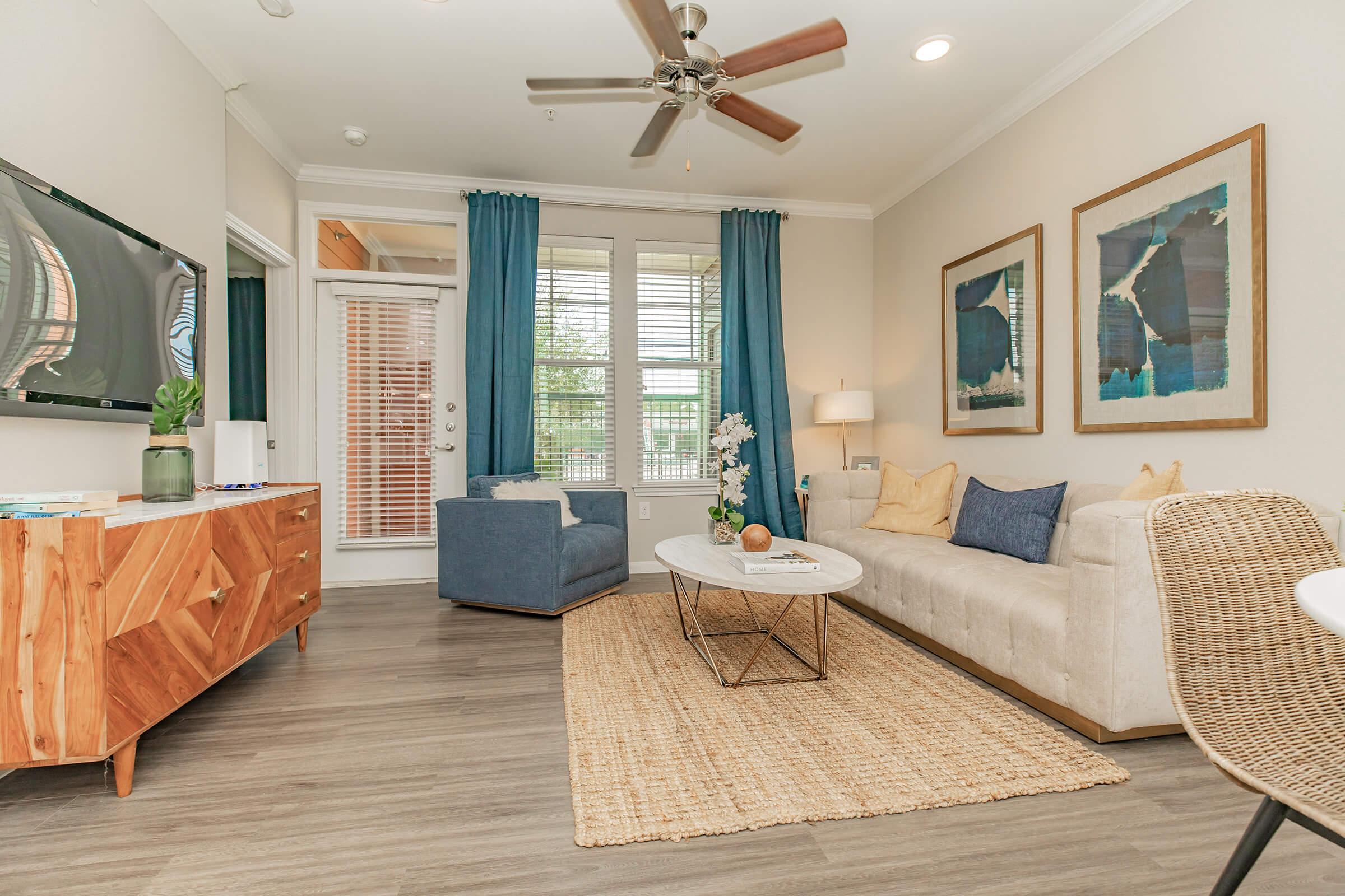 A cozy living room featuring a beige sofa and a blue armchair. There's a round coffee table on a woven rug, with a decorative plant. A wooden TV stand holds a flat-screen TV. Large windows are dressed with blue curtains, allowing natural light. The walls display abstract art, enhancing the modern aesthetic.