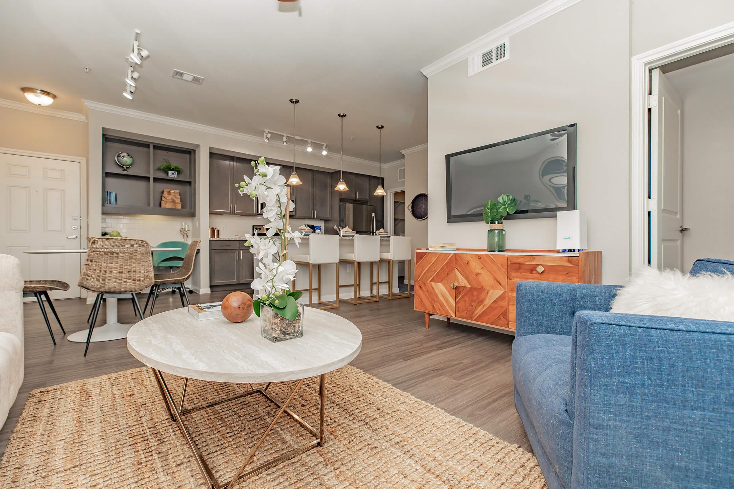 A modern living room featuring a round coffee table with a marble top, a blue sofa, and a wooden sideboard. The space has a cozy decor with a woven rug, plants, and stylish lighting. In the background, there is a kitchen with gray cabinets and bar stools, creating a welcoming open-concept atmosphere.