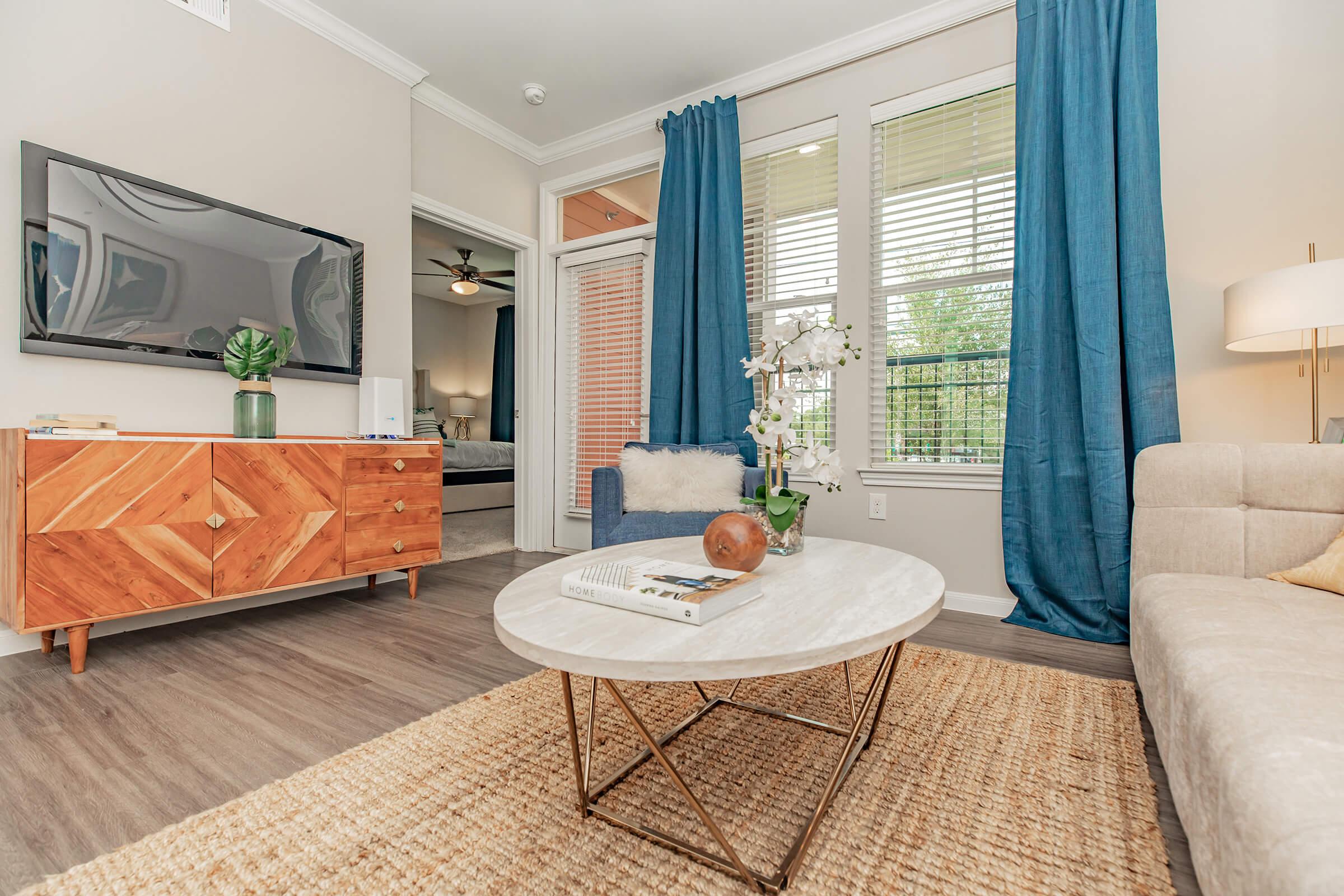 A cozy living room featuring a light-colored sofa, a round marble coffee table, and a wooden cabinet with a geometric design. Decorative plants and a vase of flowers add a touch of nature, while blue curtains frame a window, letting in natural light. A television is mounted on the wall, enhancing the modern decor.