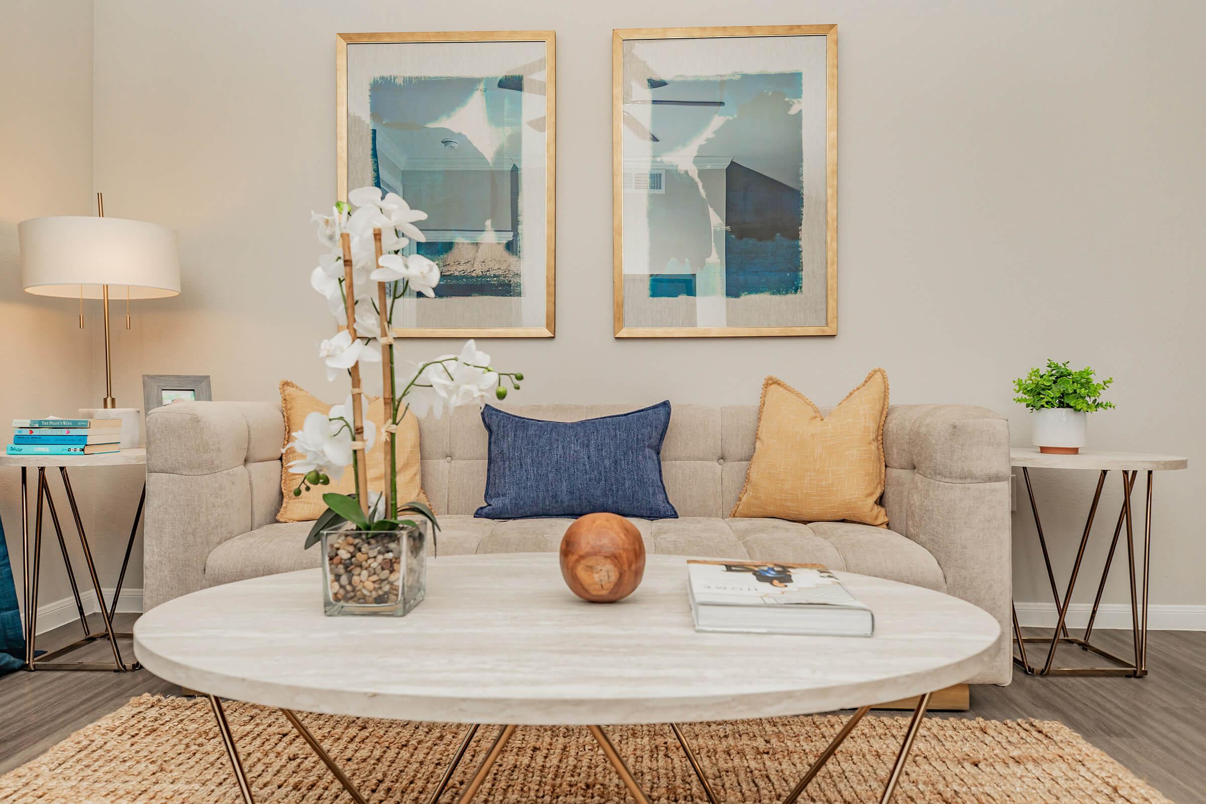 A cozy living room scene featuring a light gray sofa with decorative pillows, a round coffee table with a book and a decorative wooden bowl, and a plant. The backdrop includes two framed artworks and a lamp, creating a welcoming atmosphere. A vase with white orchids adds a touch of elegance.