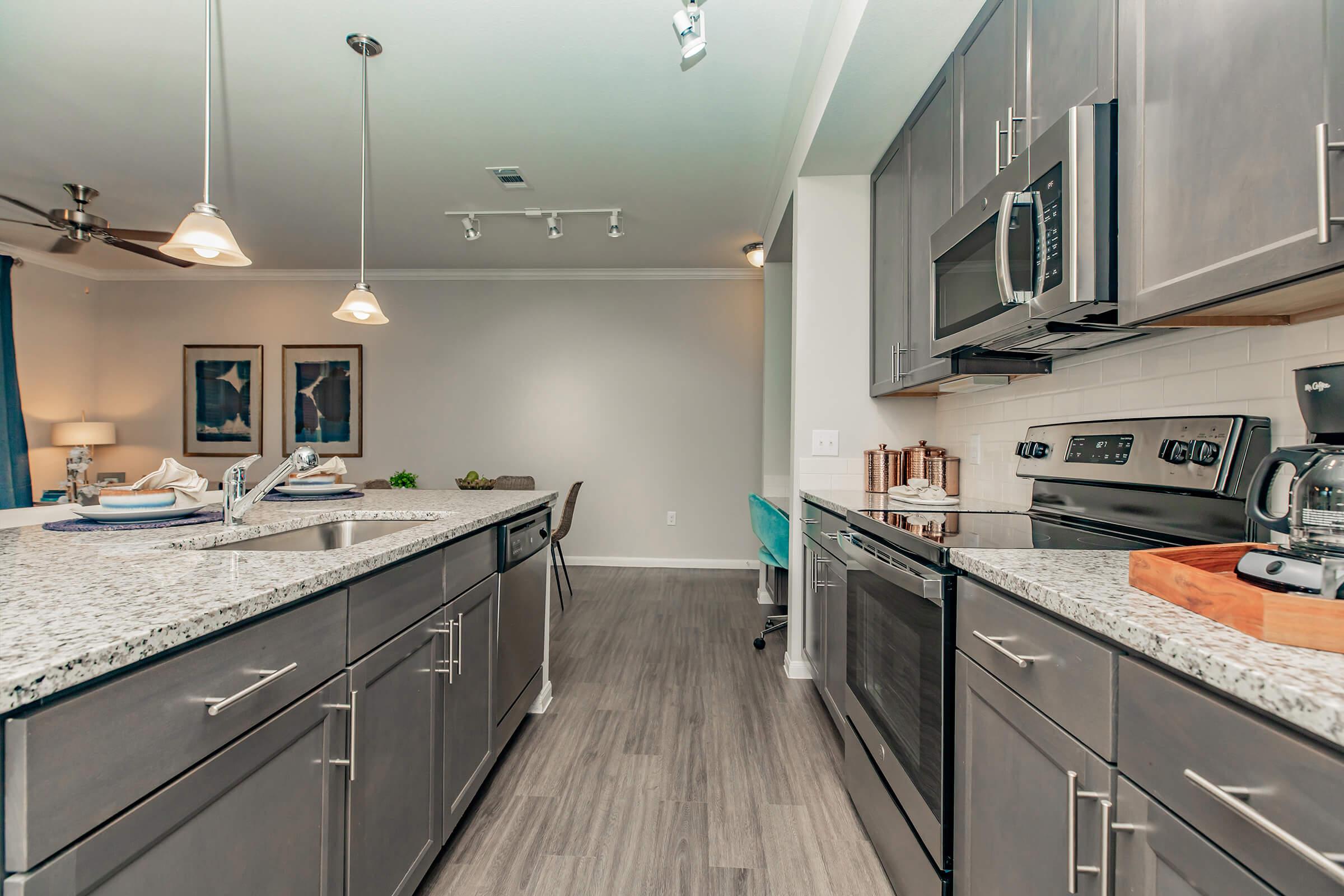 Modern kitchen featuring granite countertops, stainless steel appliances, and ample cabinet space. The design includes a large island with a sink, pendant lighting, and an open layout connecting to a living area with neutral-colored walls and minimal decor.