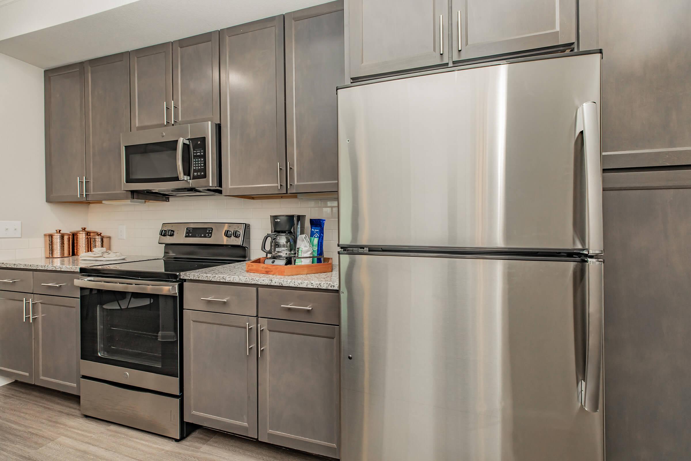 Modern kitchen featuring stainless steel refrigerator, oven, and microwave. Dark wood cabinets line the walls, and a granite countertop is visible. There is a coffee maker on the counter, along with several copper containers, creating a sleek and functional cooking space.
