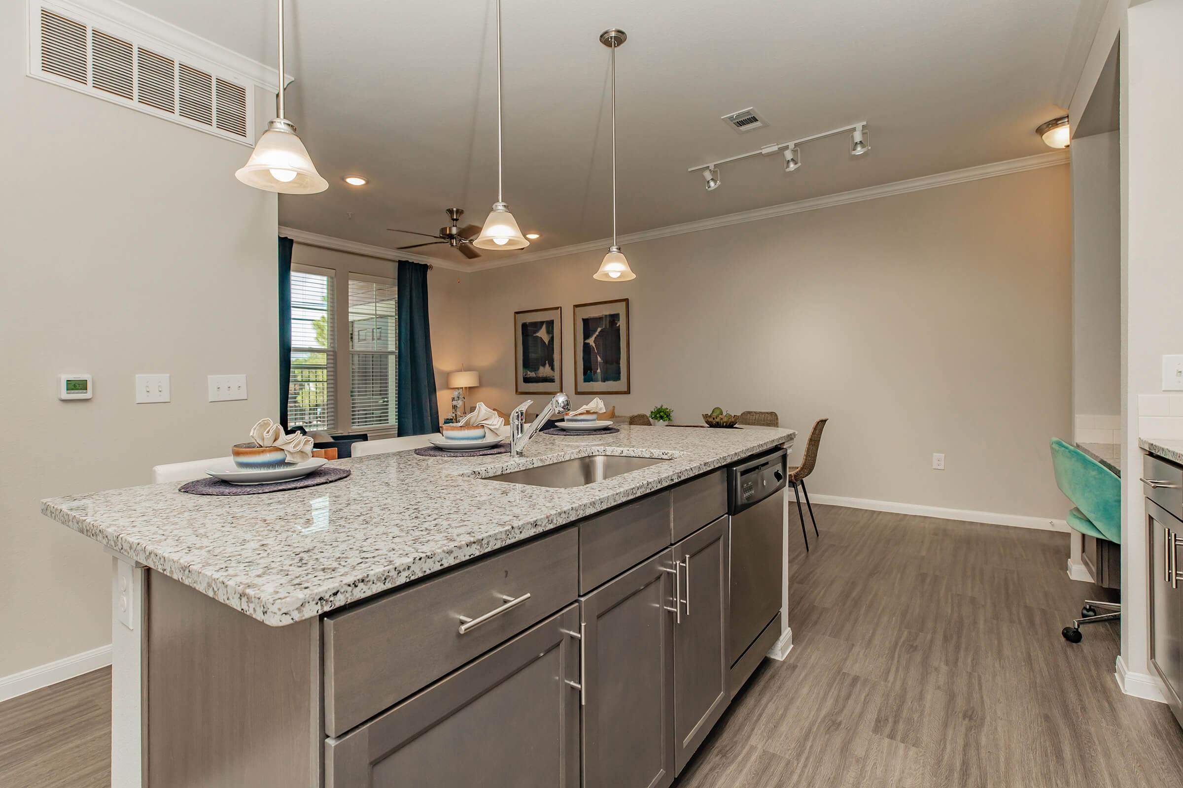 A modern kitchen featuring a large granite island with bar seating, stainless steel appliances, and pendant lighting. The room has neutral-colored walls and wooden flooring, adorned with blue curtains. In the background, there are two framed artworks and a cozy seating area with a chair and a table.