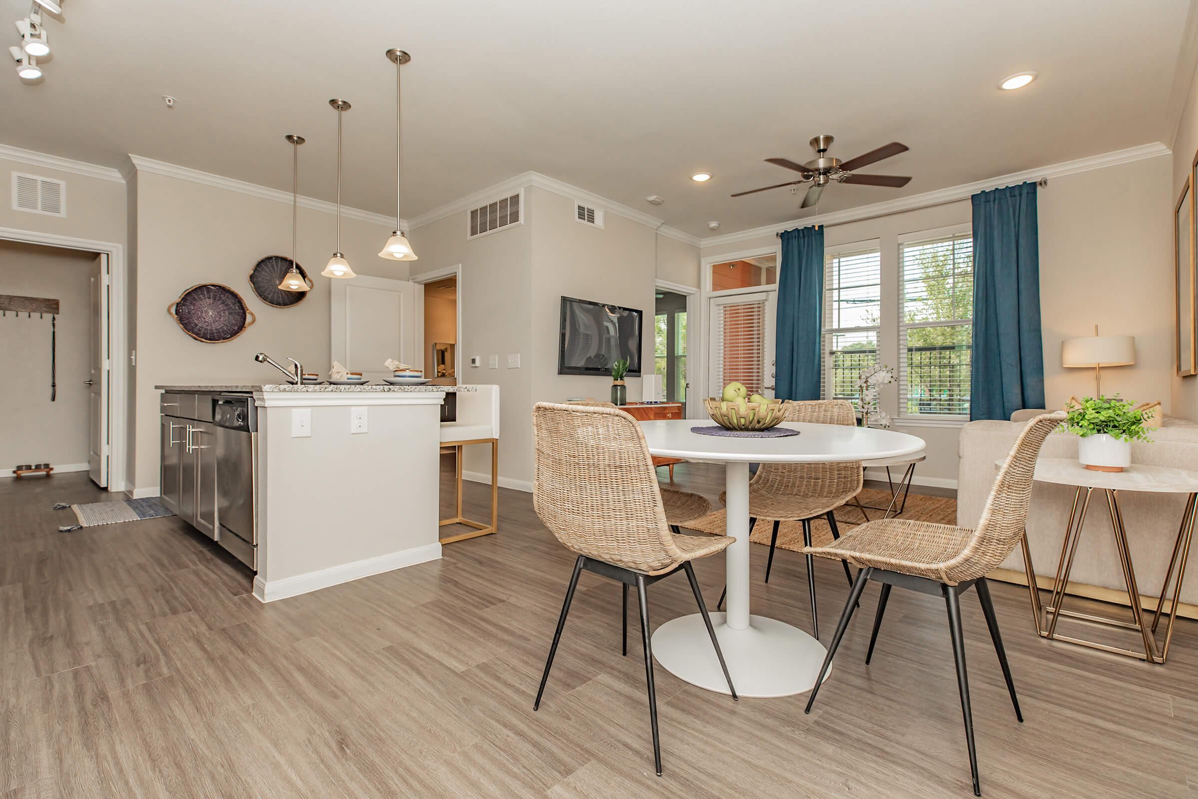 Modern kitchen and dining area featuring a round table with four woven chairs, a sleek kitchen island with bar stools, and a cozy living space with a large window. Decor includes light fixtures, plants, and soft curtains, creating a bright and inviting atmosphere.