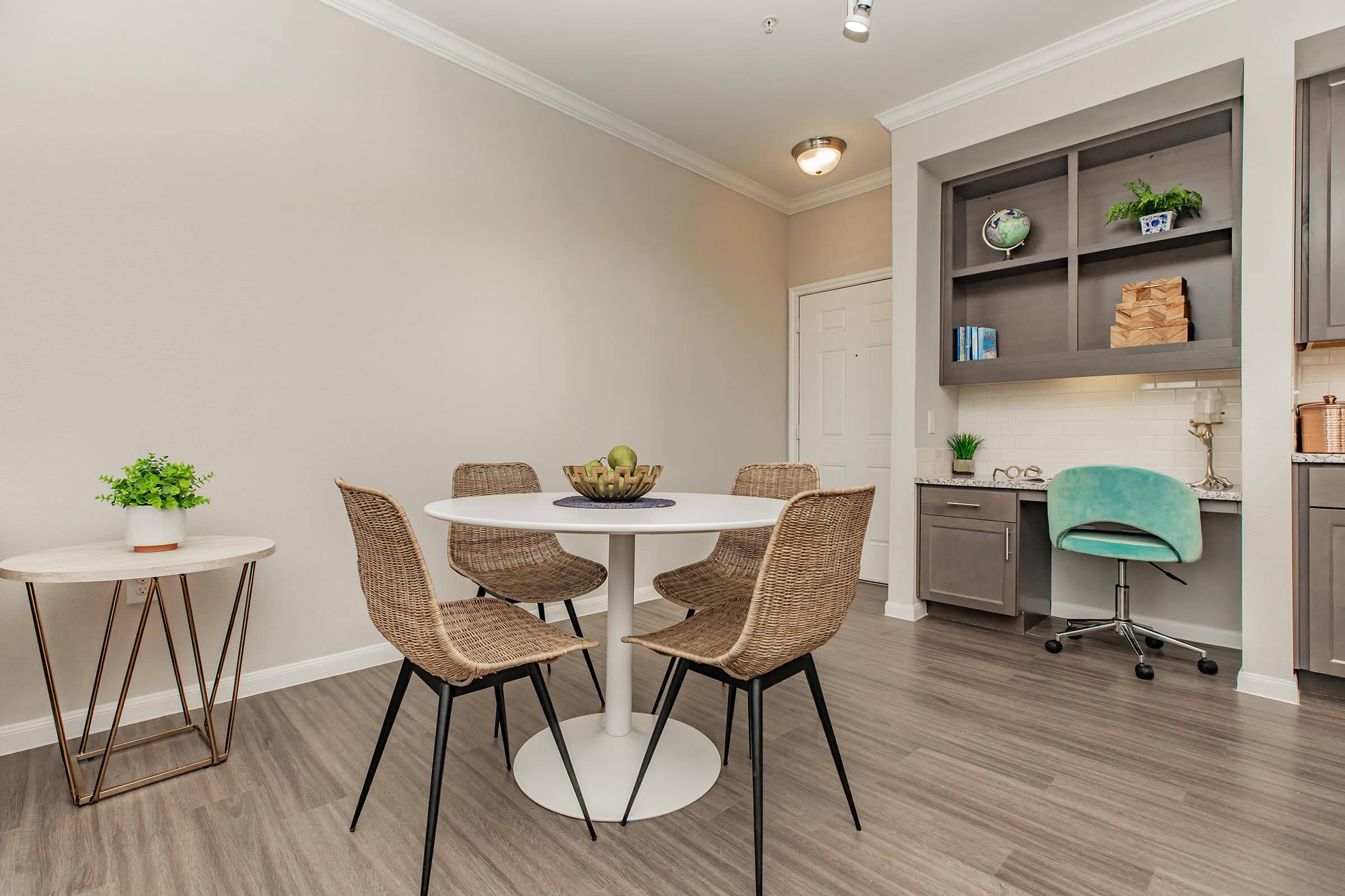 A modern dining area featuring a round white table surrounded by four woven chairs. A small side table with a potted plant is nearby. The background shows a kitchen space with grey cabinetry, a decorative shelf, and a turquoise chair at a small desk. The floor is light-colored wood.