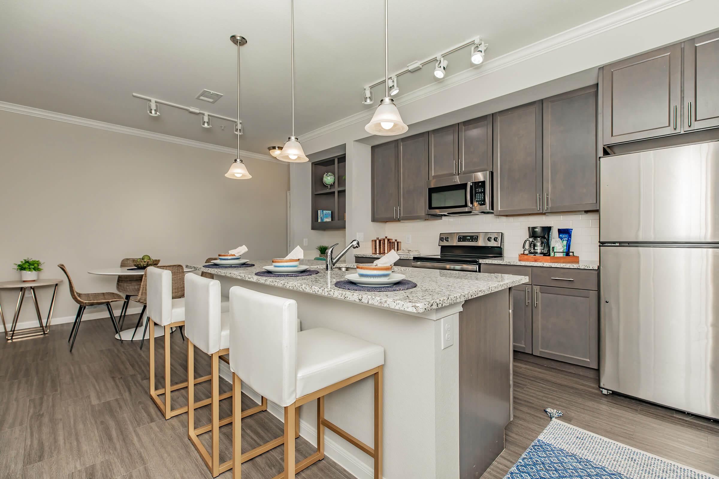 Contemporary kitchen featuring a large island with white bar stools, gray cabinetry, stainless steel appliances, and pendant lighting. A dining area with a small table and chairs is visible in the background. The space is well-lit and has a polished, modern design, showcasing an open-concept layout.